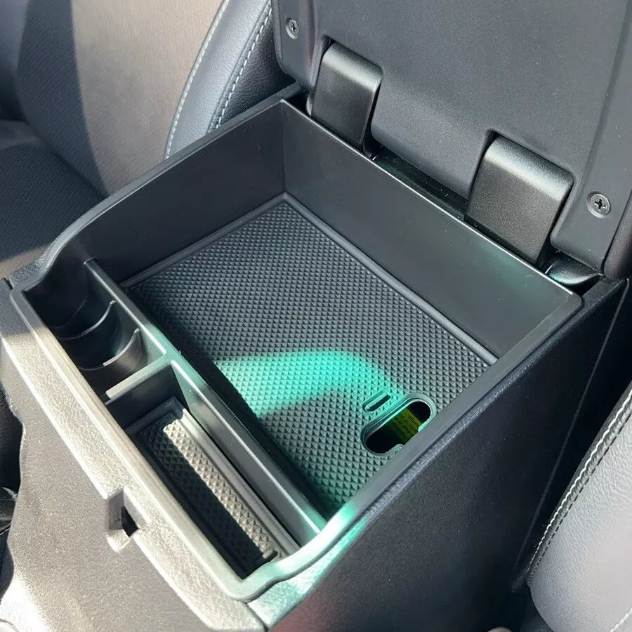 A close-up view of an aftermarket organizer tray installed inside the open center console armrest of a vehicle. The black plastic tray creates multiple compartments, including a large main section with a textured black anti-slip rubber mat and smaller sections for coins or phones. The surrounding interior of the car features black upholstery with white stitching.
