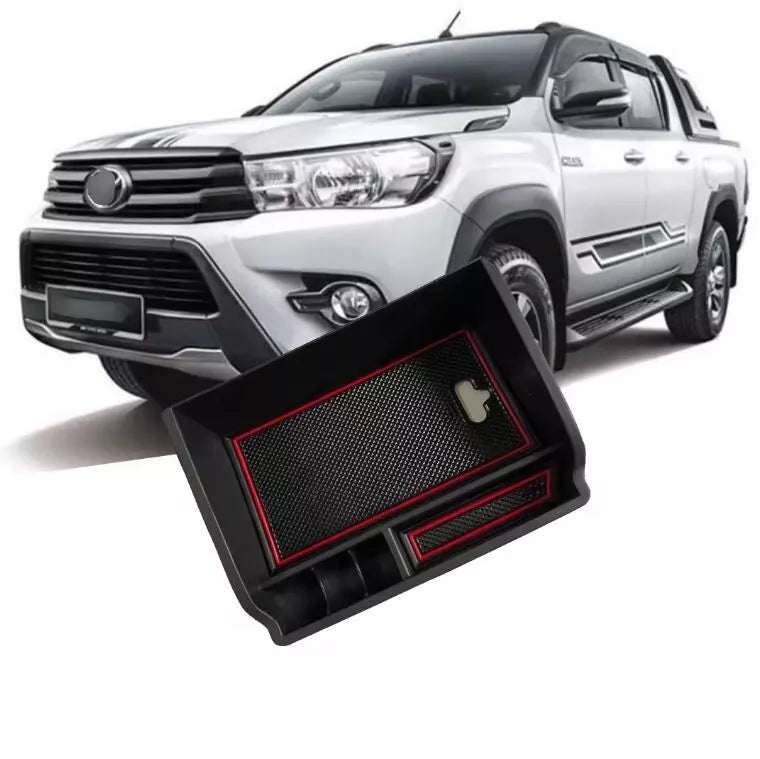 A promotional image featuring a black plastic organizer tray, designed to fit into a vehicle's center console, floating in the foreground. The tray has red accents and compartments. In the background, a white, four-door Toyota Hilux pickup truck is visible.