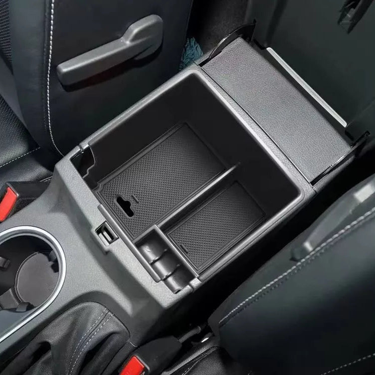 A close-up image showing a black, custom-fit organizer tray placed inside an open car center console storage compartment.
The tray has multiple divided sections, including a larger main section, a narrower section, and two small square compartments near the front. The surfaces inside the tray have a textured, rubberized, or non-slip material. A USB port is visible on the front vertical wall of the console, accessible even with the tray in place. The surrounding interior of the car features black leather se