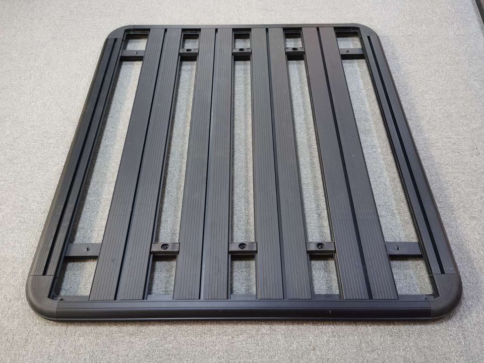 A rectangular, heavy-duty flat platform roof rack for a vehicle, shown against a grey carpet background. The rack is black, likely made of aluminum, and features several long, parallel load-bearing slats with a low-profile outer frame.