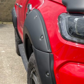 A tight close-up view of the front wheel arch and surrounding body panel of a bright red pickup truck. The truck is equipped with a wide, matte black bolt-on style fender flare that covers the wheel well and extends down to the bumper. The flare has visible silver or metal-look rivets/bolts. A side step is partially visible beneath the door further back.