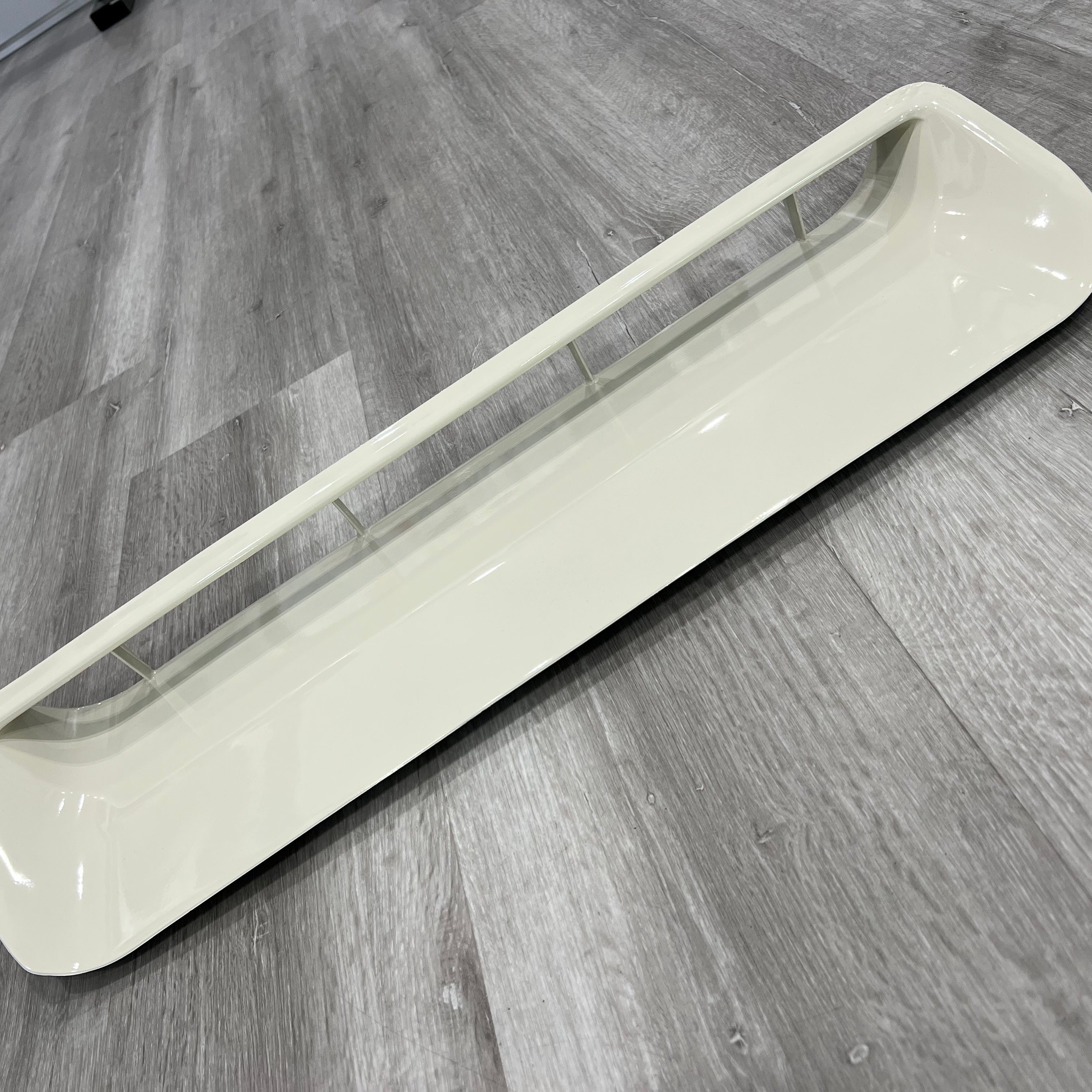 A close-up, diagonal view of a single, long, rectangular automotive hood scoop or air vent, painted in a glossy light beige or cream color. The central opening features a single horizontal bar supported by several small vertical struts. The scoop is resting on a gray wood grain floor.