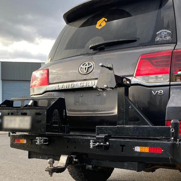 A rear view of a dark gray Toyota Land Cruiser 200 Series SUV, parked on asphalt under a cloudy sky. The vehicle is equipped with a heavy-duty, matte black steel rear bumper and dual swing-out carrier system.
The bumper assembly features:
A storage basket/jerry can holder on the left side, designed to swing out.
A spare tire carrier mounting post on the right side, also designed to swing out.
Integrated amber and red lights on the lower corners of the bumper.