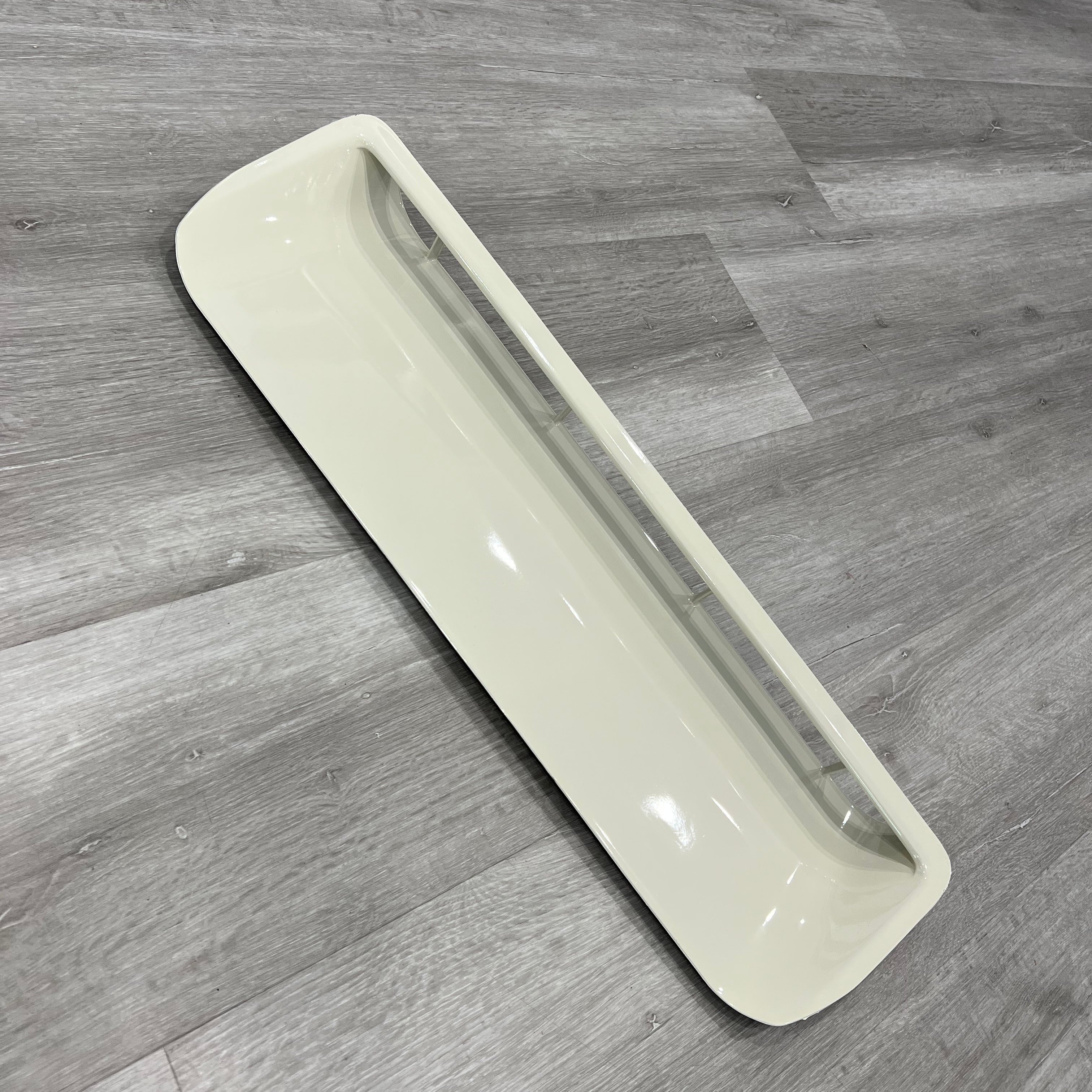 A rectangular, off-white or beige plastic component, which appears to be the cover or trim for an air vent or duct opening. It has rounded corners and a smooth, glossy surface, lying on a gray wood-look floor.