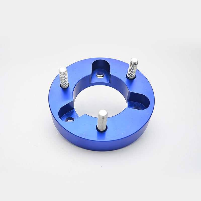 A top-down, close-up product image of a single, thick, blue anodized aluminum wheel spacer or leveling lift spacer. The round spacer features cutouts around the center opening and has three silver studs extending upwards, ready for mounting. The surface is reflective, indicating a quality finish.