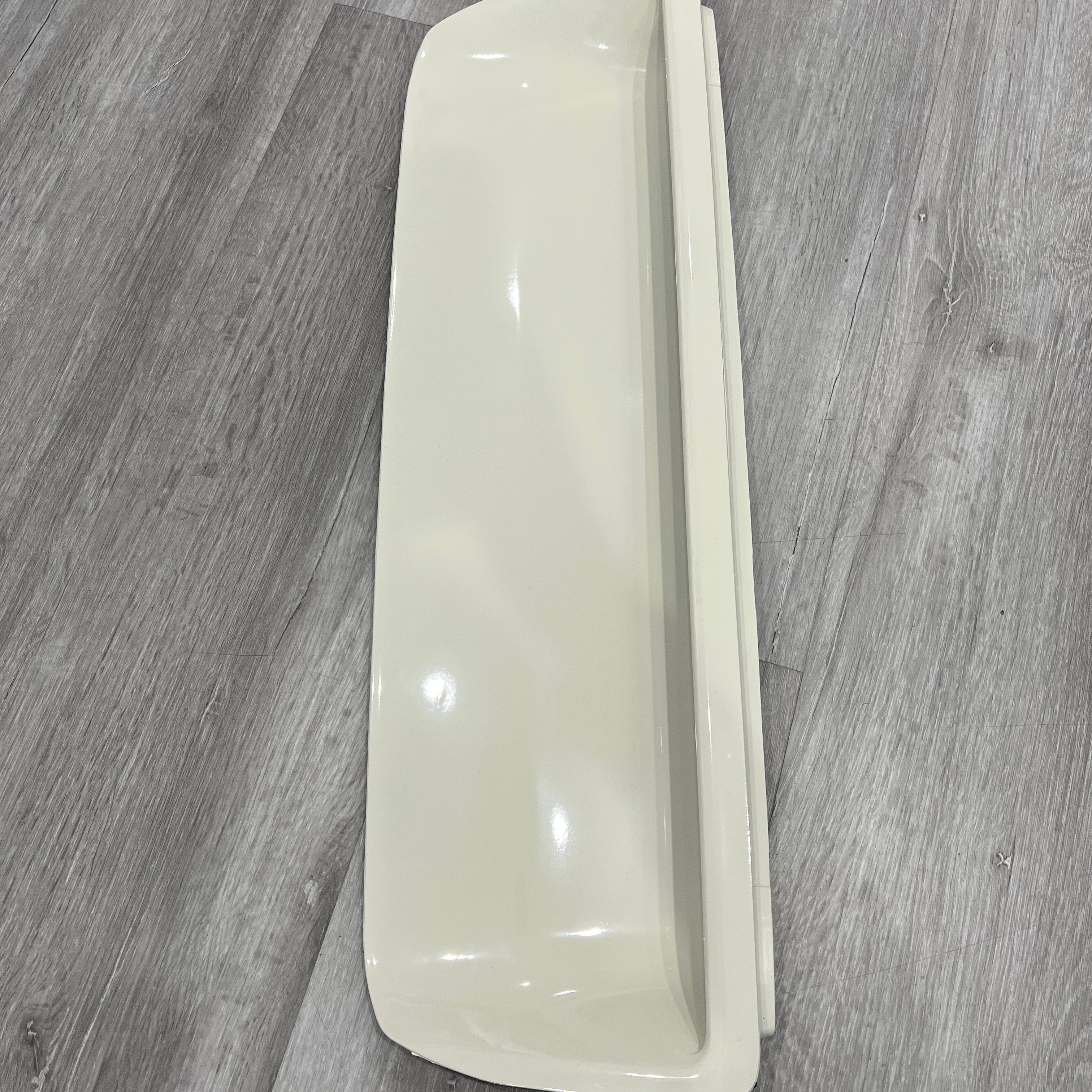 A vertical, close-up view of a single, long, rectangular automotive fender vent or air intake scoop, painted in a glossy light beige or cream color. The scoop has a deep recess or opening running vertically along its right side. It is resting on a gray wood grain floor.
