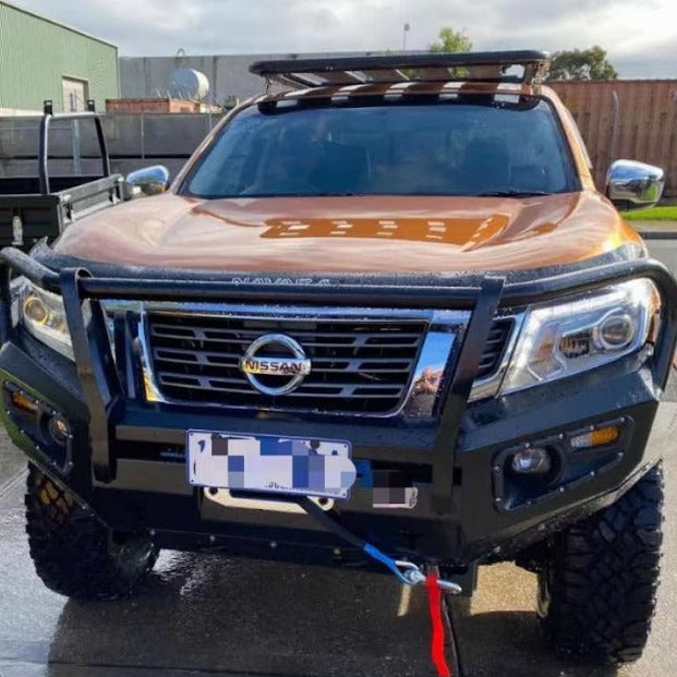 A front-view photo of an orange or copper-colored Nissan Navara pickup truck, modified with off-road accessories. It features a heavy-duty black steel bull bar and bumper, a flat black roof rack, and large all-terrain tires. A blue and red recovery strap is attached to the winch fairlead opening. The vehicle is parked outdoors in what appears to be an industrial area, with part of a utility truck visible on the left.