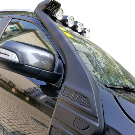 A low-angle, close-up photograph of the front passenger side of a black pickup truck with off-road modifications. The most prominent features are the large, black plastic snorkel mounted on the A-pillar and fender, featuring a honeycomb grille at the air intake. The snorkel head is topped with a row of four round LED auxiliary lights. The reflection of a building is visible in the side window and a digital checkered pattern fills the background.