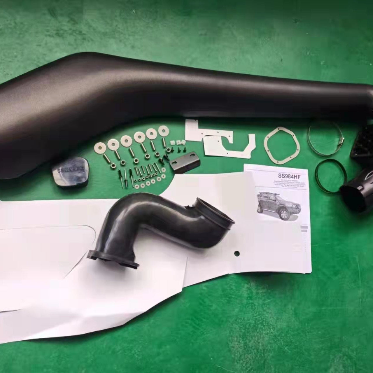 A flat lay photo of a complete black automotive snorkel kit (SS984HF), laid out on a dark green surface. The major components are the long, curved main snorkel pipe and a black rubber or plastic elbow air intake hose. The accompanying parts include mounting hardware (bolts, washers, nuts, and a small black bracket), a paper instruction manual showing an image of a 4x4 vehicle, a large white paper installation template, and various seals and clamps.