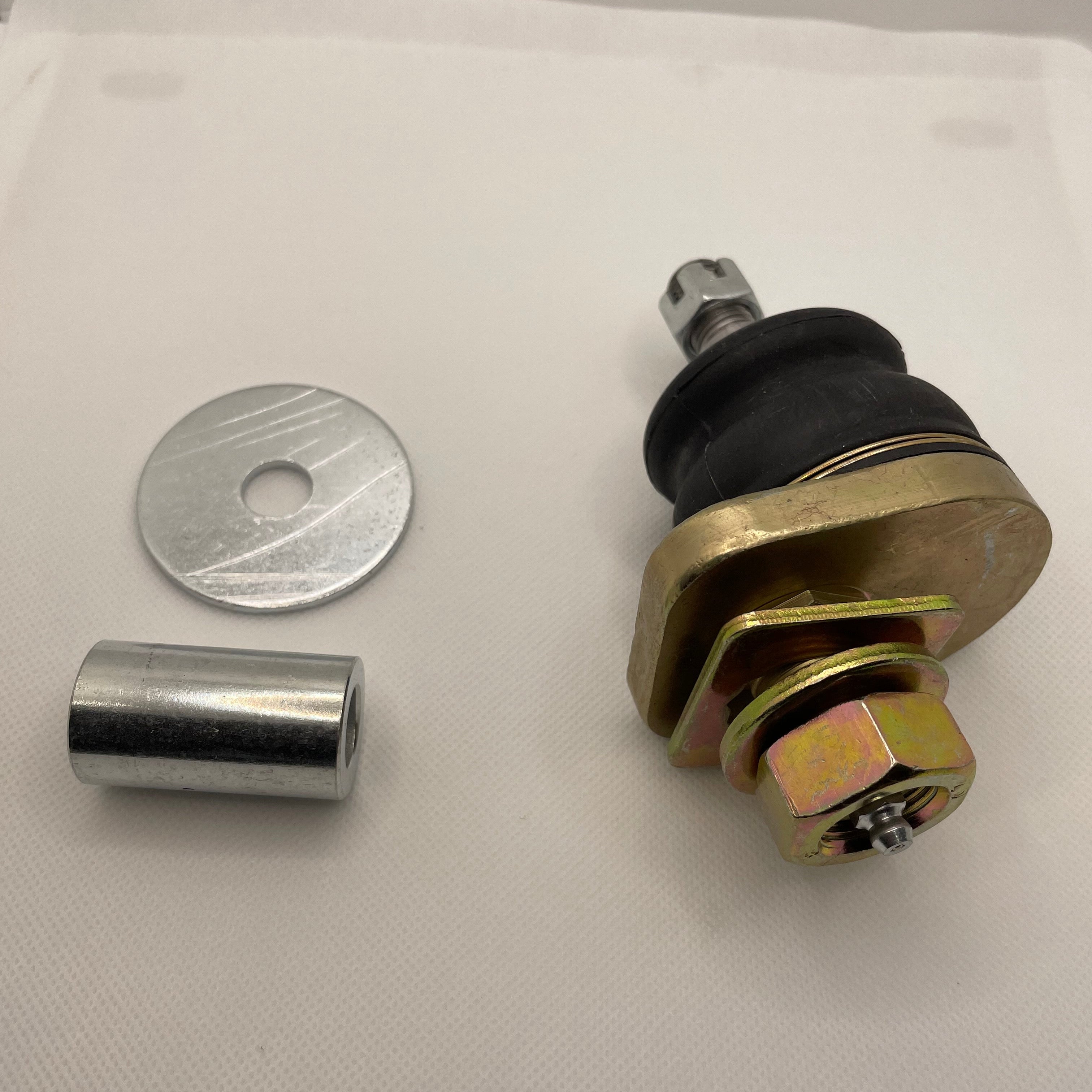 A product image showing a heavy-duty ball joint assembly for a vehicle suspension, along with two pieces of separate hardware, placed on a white surface. The ball joint is assembled with a black rubber boot, gold-colored mounting plate, and is secured by a large gold-colored nut at the bottom and a castellated nut and cotter pin on the threaded stud at the top. To the left, there is a large silver washer and a shiny silver metal sleeve or bushing insert.