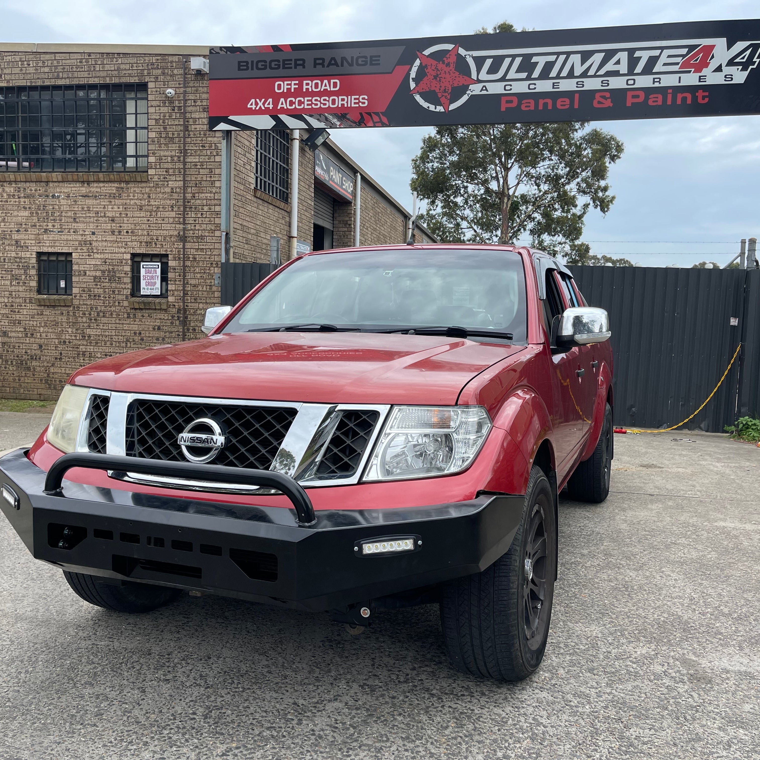 A red Nissan Navara (D40 generation) pickup truck is parked outside a business called "Ultimate 4x4 Panel & Paint." The truck is fitted with a black, heavy-duty aftermarket steel bull bar/bumper that includes a single black tubular hoop and an integrated horizontal LED light bar on the driver's side. The background shows a brick building with a large red and black sign above, and a dark fence to the right.