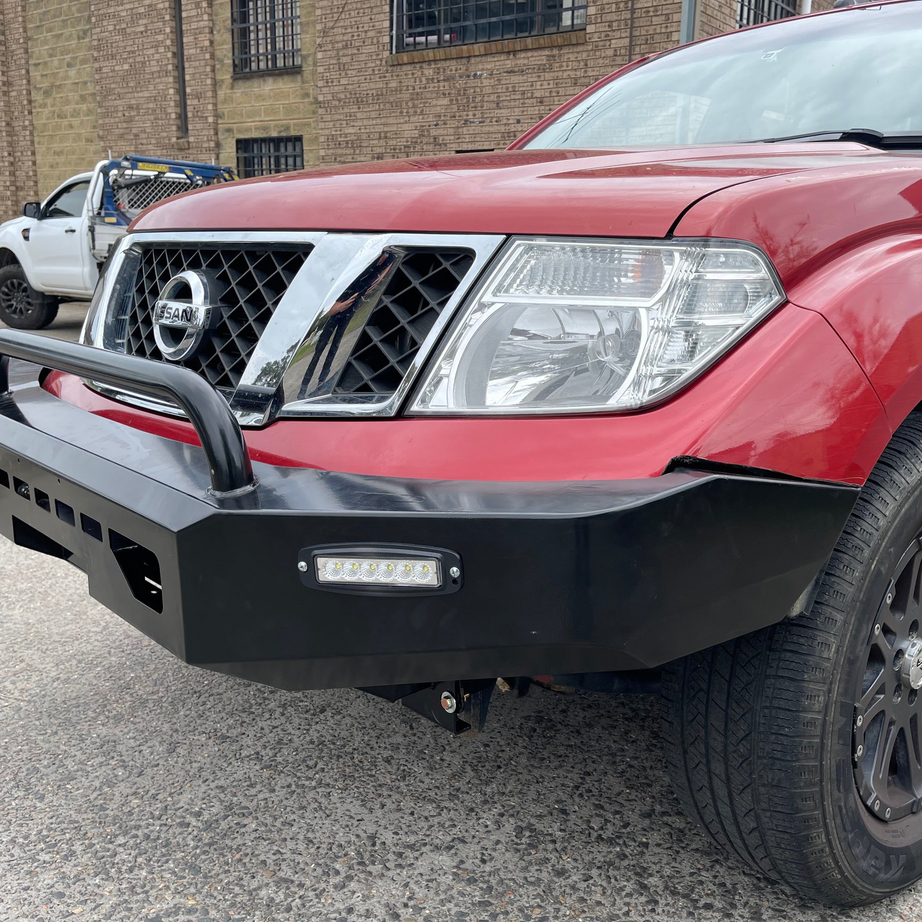 A close-up, low-angle shot of the front of a red Nissan Navara or Frontier (D40 generation) pickup truck. The truck is fitted with a black, angular heavy-duty aftermarket steel bull bar/bumper. The bumper features a single black tubular hoop, an integrated horizontal LED light bar on the passenger side, and a cutout for a winch fairlead. In the background, a white utility truck is partially visible parked outside a brick building.