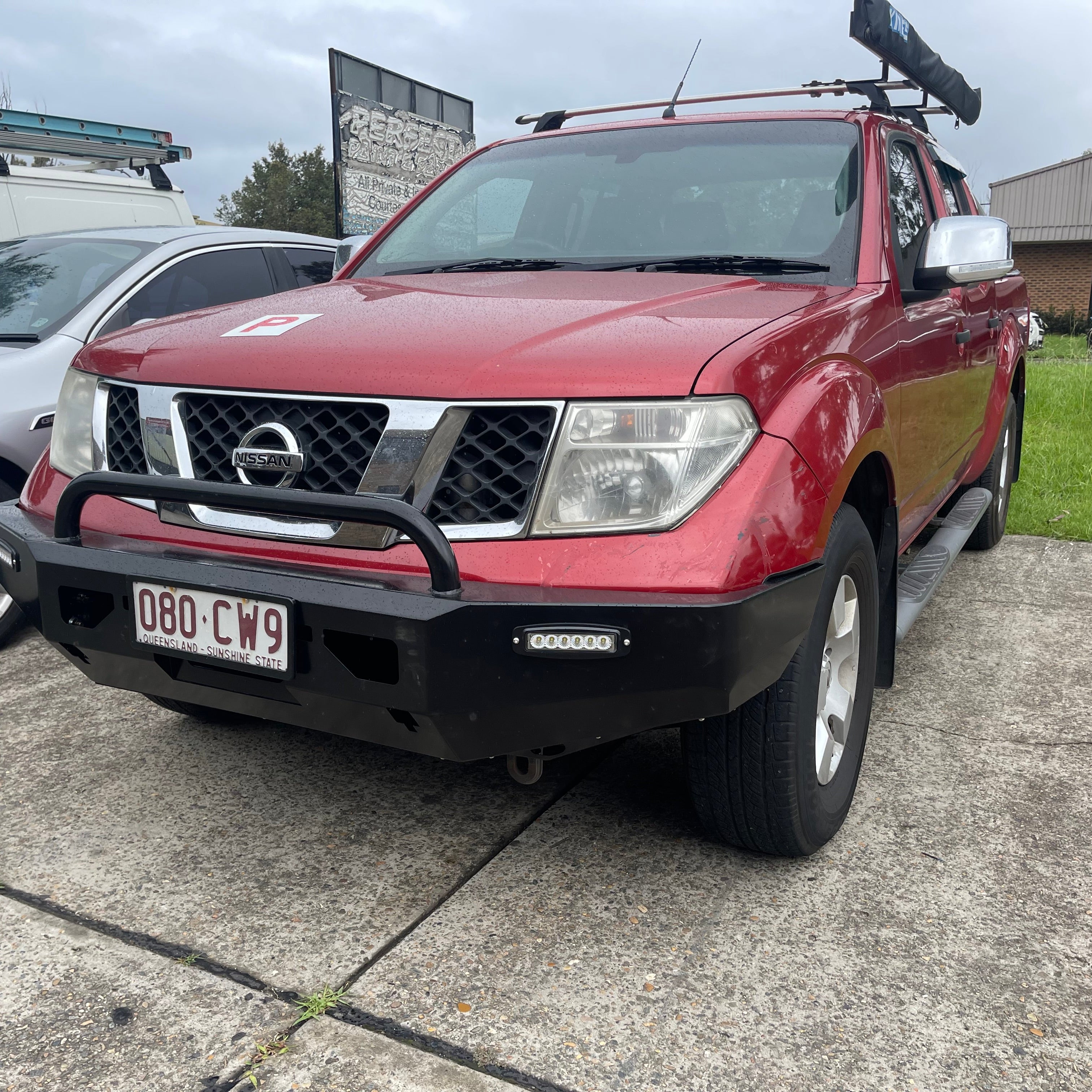 A red Nissan Navara (D40 generation) pickup truck is shown from a front-quarter view, parked on a concrete surface. It is equipped with a black, heavy-duty aftermarket steel bull bar/bumper that includes a single black tubular hoop and an integrated horizontal LED light bar on the driver's side. The truck features silver mirror caps, roof racks carrying a black awning or tube, and a red 'P' plate (indicating a probationary driver) on the hood.