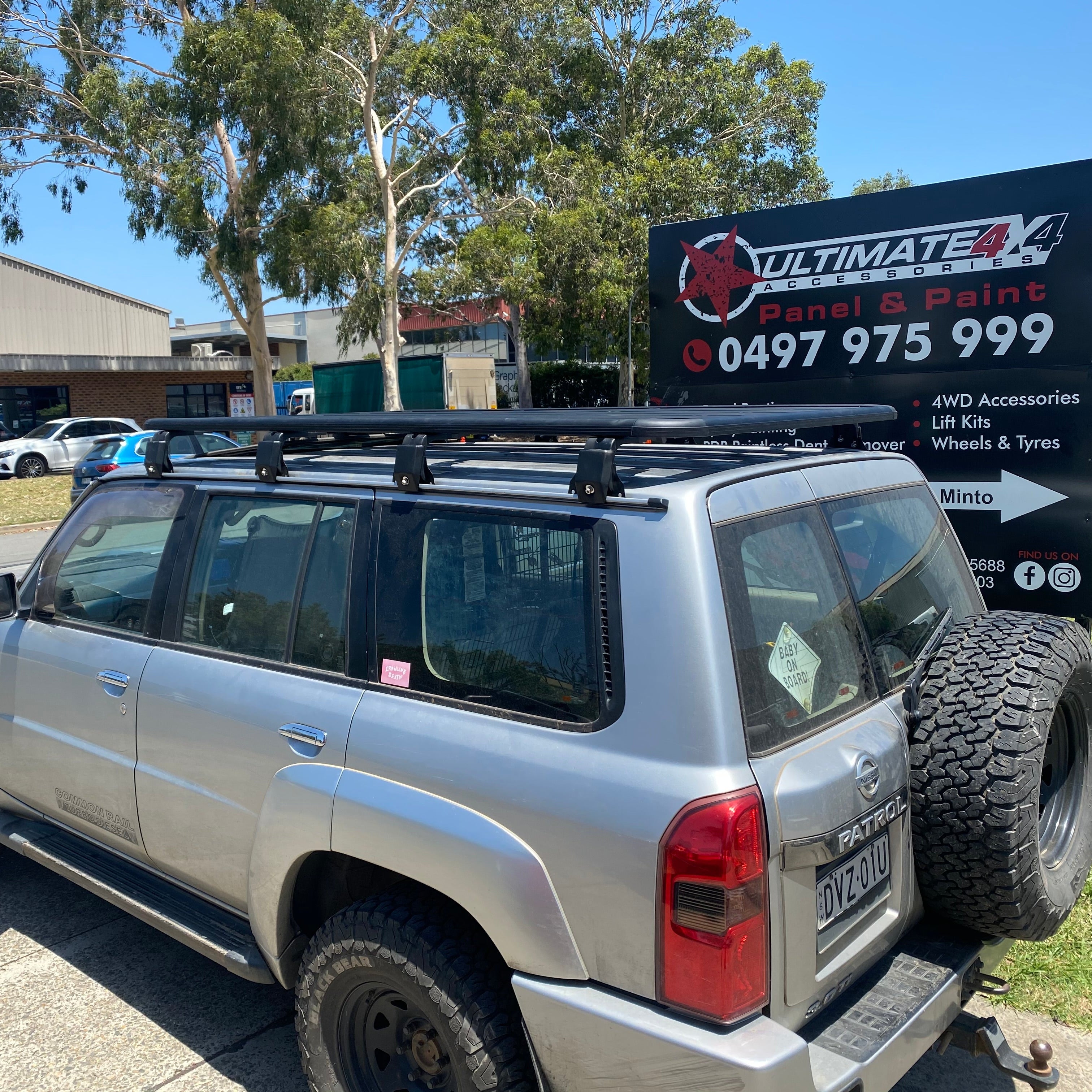 A three-quarter rear view of a silver Nissan Patrol SUV with a black, heavy-duty roof rack installed. The vehicle has a spare off-road tire mounted on the back door and off-road tires on black wheels. The SUV is parked outdoors in front of a large black sign for "ULTIMATE 4X4 ACCESSORIES Panel & Paint" with red and white text, under a sunny sky with trees in the background.