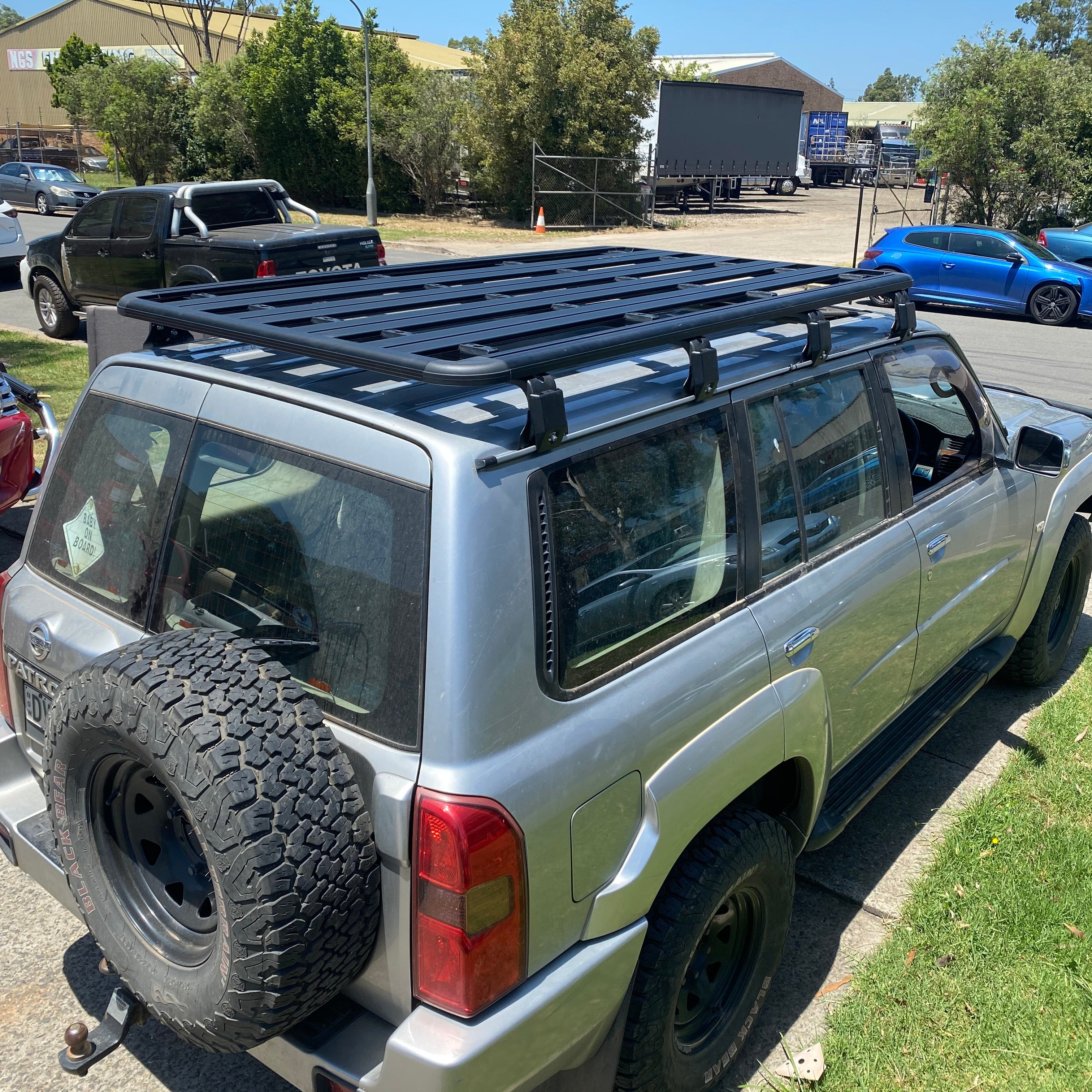 A rear three-quarter view of a silver Nissan Patrol SUV with an extra-large, black, slatted platform-style roof rack installed on the top. The SUV has an off-road tire mounted on the rear door and is parked on grass and pavement in an industrial or outdoor storage area on a sunny day. Other vehicles, including a black pickup truck and a blue hatchback, are visible in the background.
