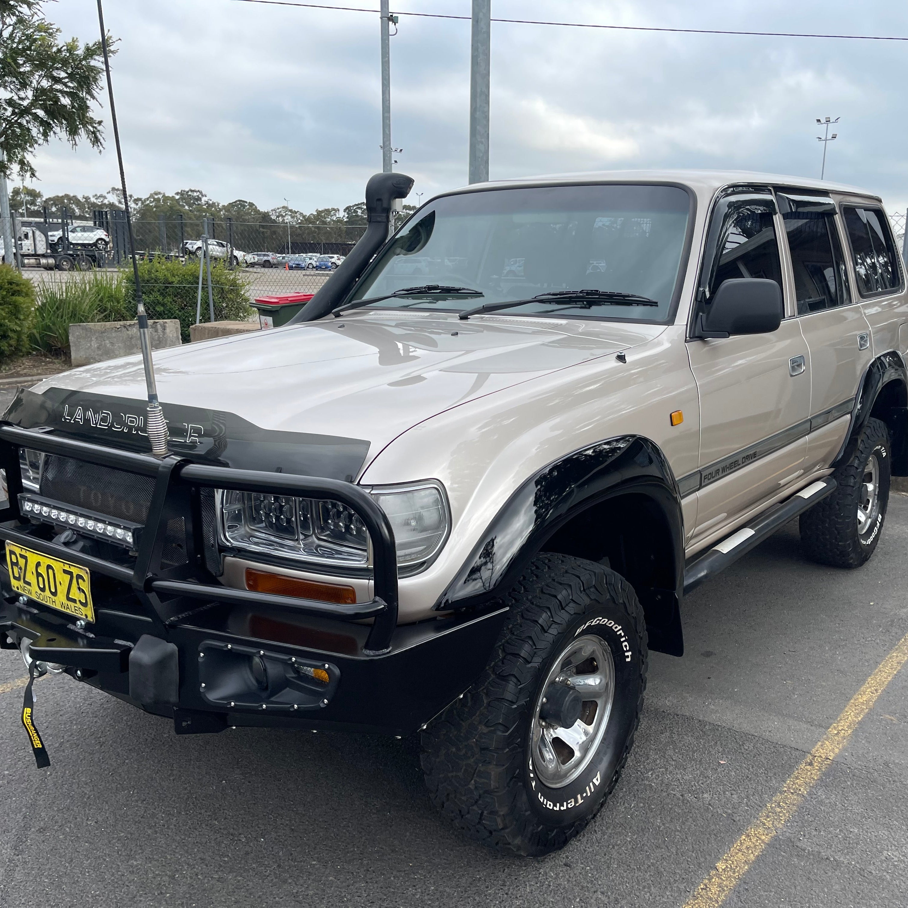 A modified champagne gold/beige Toyota Land Cruiser 80 series (FJ80/HZJ80) parked outdoors in a lot on an overcast day. The vehicle is equipped with a black steel bull bar holding an LED light bar and a radio aerial, a black snorkel, black fender flares, and BFGoodrich All-Terrain T/A tires on chrome wheels. It has a New South Wales, Australia license plate.