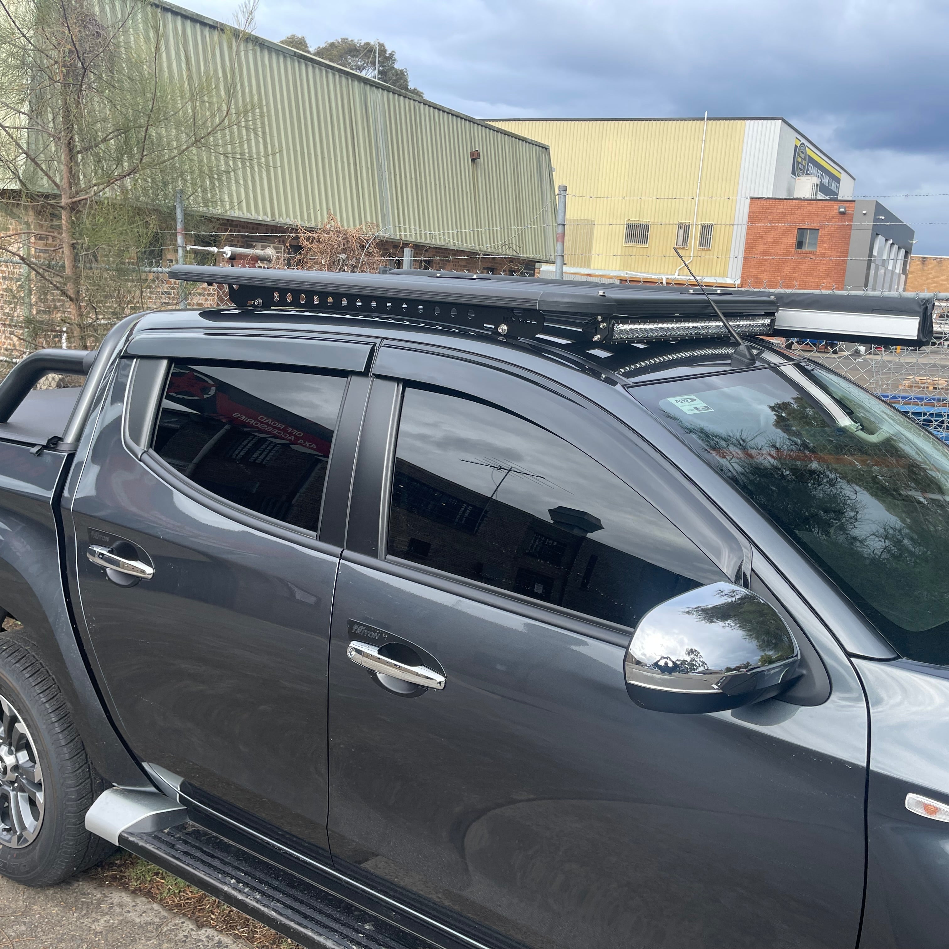 A slightly angled side view of a dark gray pickup truck (likely a double cab) featuring a low-profile, flat black roof rack platform over the cab. The rack is fitted with a large LED light bar mounted on its front edge. The truck has tinted windows, weather deflectors, chrome door handles and side mirrors, and silver side steps. The background shows an outdoor setting with large industrial or commercial buildings with corrugated metal siding under a cloudy sky.