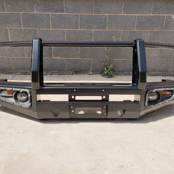 A standalone black or dark gray steel bull bar or replacement bumper for an off-road vehicle. It features a triple-loop design with upright central posts and integrated light housings on either side for fog or indicator lights. The bumper includes a central opening for a winch fairlead and a mounting space for a license plate. The bull bar is photographed against a background of a dark gray brick wall and a concrete floor.