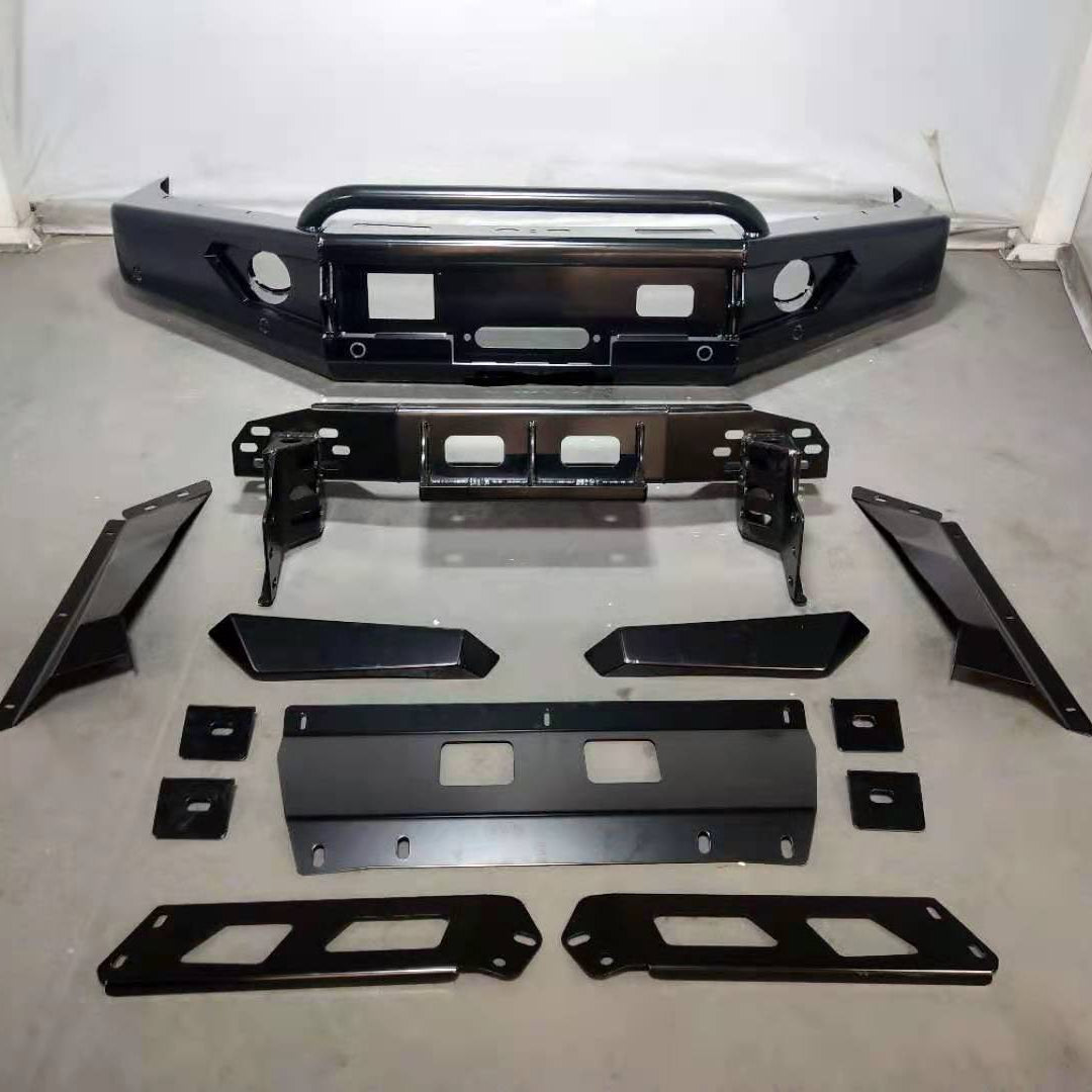 A complete, glossy black aftermarket steel front bumper kit for an off-road vehicle, laid out on a gray floor against a light gray wall. The main bumper assembly, positioned at the top, is an angular, heavy-duty design with a tubular top hoop, cutouts for two fog lights, and a central opening for a winch fairlead. Below the bumper, the kit components are arranged, including a large winch mounting plate, various mounting brackets, smaller plates, and two side wing.