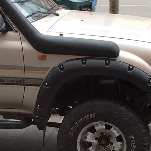 A close-up shot of the front passenger side of a tan Toyota Land Cruiser (likely a 70 or 80 series, judging by the shape and "DIESEL DRIVE" badge). The vehicle is equipped with several off-road modifications: a black snorkel running up the side of the hood and A-pillar, and black bolt-on fender flares covering the wheel well. A large off-road tire is visible, partially obscured by the flare. The word "DIESEL DRIVE" is faintly visible on the side panel near the door.