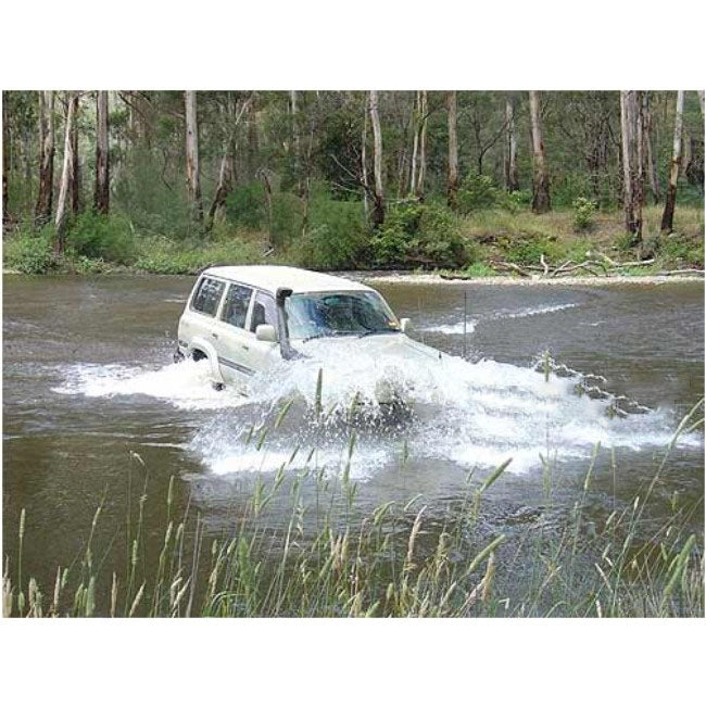 A dynamic outdoor photograph showing a white four-wheel-drive SUV (appearing to be an older model Toyota Land Cruiser 80 series) driving through a shallow river or creek, creating a large splash of water that obscures the front bumper. The vehicle is fitted with a black snorkel on the driver's side A-pillar. The background shows trees and dense vegetation.