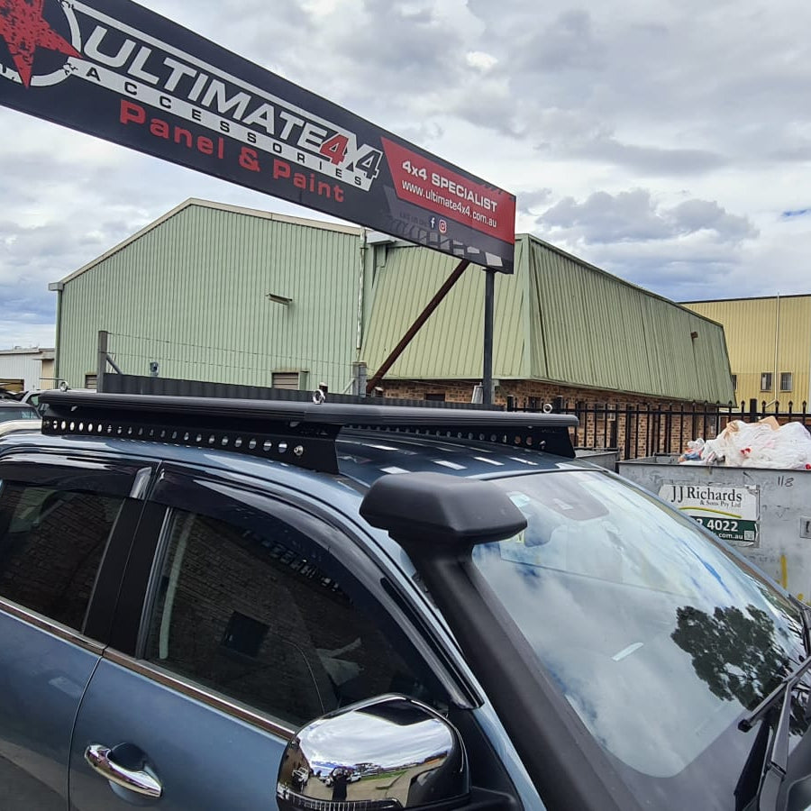 A cropped photo focusing on the front and roof of a metallic blue pickup truck, showing two 4x4 accessories: a black snorkel mounted on the passenger side A-pillar and a low-profile black platform roof rack with a perimeter frame and slats. A large red and black sign for "ULTIMATE 4X4 ACCESSORIES Panel & Paint" is visible above the truck, with industrial buildings in the background under an overcast sky.