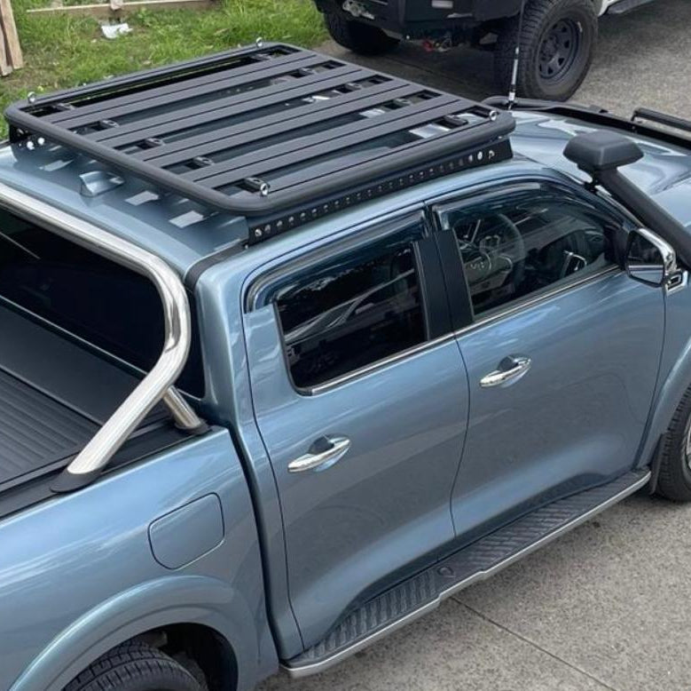 A high-angle photo focusing on the cab and bed of a metallic blue pickup truck, possibly a Nissan Navara, equipped with 4x4 accessories. Key features visible are a large black platform roof rack mounted over the cab, a black snorkel on the passenger side, a silver sports bar over the bed, a black roller shutter covering the bed, and side steps. Another vehicle is partially visible in the background.