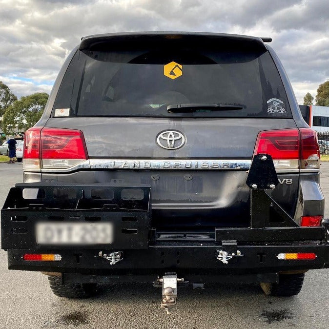 A rear view of a dark gray Toyota Land Cruiser 200 Series SUV, parked outdoors on asphalt under a cloudy sky. The vehicle is fitted with a heavy-duty, black steel rear bar featuring a dual swing-out carrier system in the closed and latched position.
The features of the rear bar include:
A storage/jerry can carrier on the left side.
A spare tire carrier mount on the right side.
Integrated amber/red lights on the lower corners.
A receiver hitch with a tow ball assembly attached.