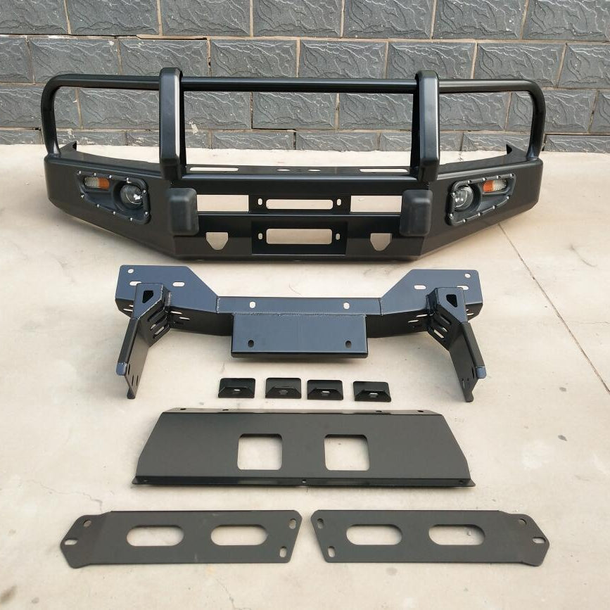 A complete, disassembled aftermarket steel bull bar/bumper kit is laid out on a gray paved surface against a brick wall background. The main bumper assembly is at the top, finished in matte black, featuring a multi-hoop upper guard, integrated circular fog lights and amber turn signals in the wings, a winch fairlead opening in the center, and two black rubber pads/bumpers below the main hoops. Below the bumper are the mounting and reinforcement components, including a heavy-duty central frame bracket.