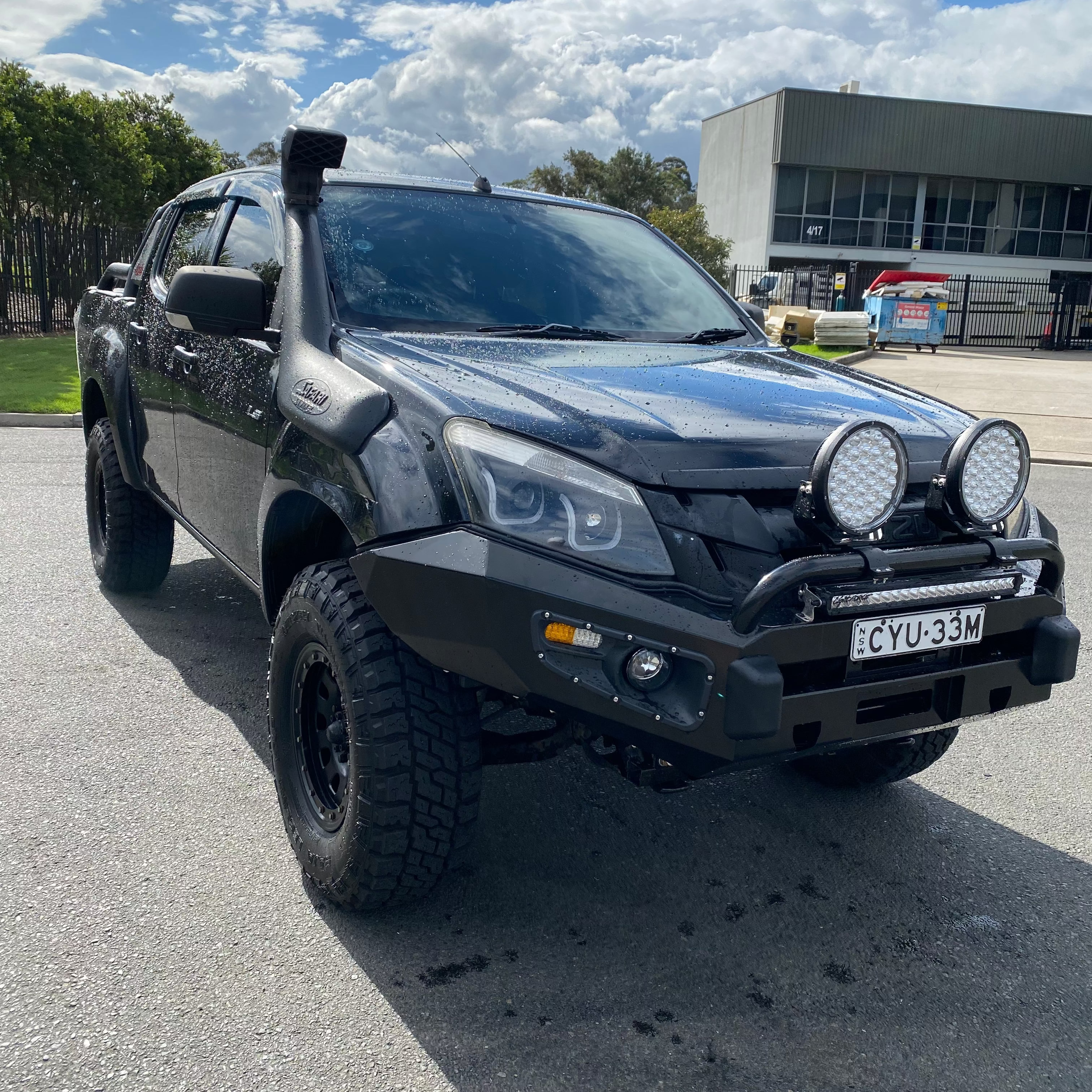 A black, heavily modified Isuzu D-Max pickup truck, seen in a front three-quarter view, is parked on asphalt in an industrial or commercial area. The truck is lifted and features a black steel bull bar, two large round LED driving lights, an LED light bar, a snorkel, and large off-road tires. The license plate reads "CYU-33M". In the background is a building and some industrial bins under a cloudy sky.