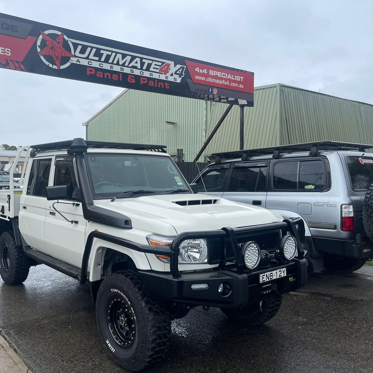 A modified white Toyota Land Cruiser 79 Series dual cab utility truck is parked outdoors on a damp day in front of a 4x4 accessories shop. The truck is heavily equipped with a black steel bull bar holding two large LED driving lights, a black snorkel, oversized off-road tires, a tray with a roll bar, and a black flat platform roof rack. The license plate reads ENB-12Y. Behind it is a silver Toyota Land Cruiser wagon also fitted with a roof rack. A sign for "ULTIMATE 4X4 ACCESSORIES Panel & Paint" is visibl