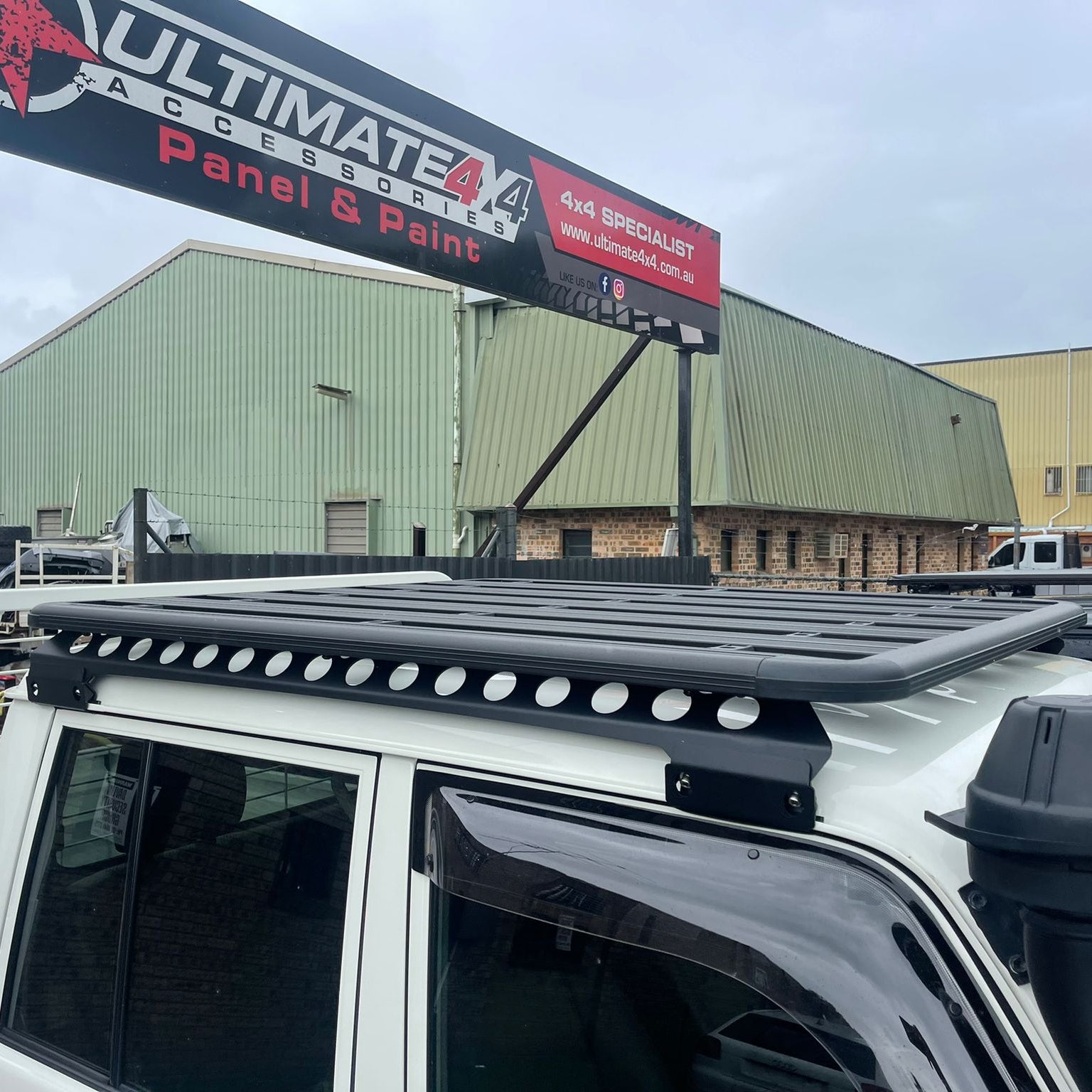A close-up view of a black, flat platform roof rack mounted on the cab of a white vehicle, likely a Toyota Land Cruiser, showing the side profile and mounting brackets. The roof rack features a unique side rail with circular cutouts. In the background, there is a large sign reading "ULTIMATE 4X4 ACCESSORIES Panel & Paint" above a couple of industrial buildings under an overcast sky.