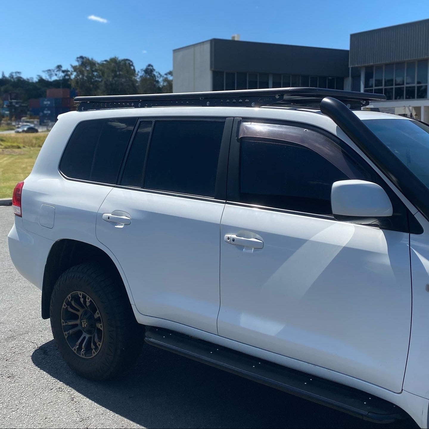 A side view of a white SUV (likely a Toyota Land Cruiser 200 Series) parked outdoors in a lot. The vehicle is equipped with several aftermarket accessories, including a black platform roof rack, a black snorkel mounted on the front right pillar, dark tinted windows, black side steps, and off-road tires with aftermarket black and bronze wheels. In the background are modern commercial buildings and a bright blue sky. The image focuses on the vehicle's profile and its overland modifications.