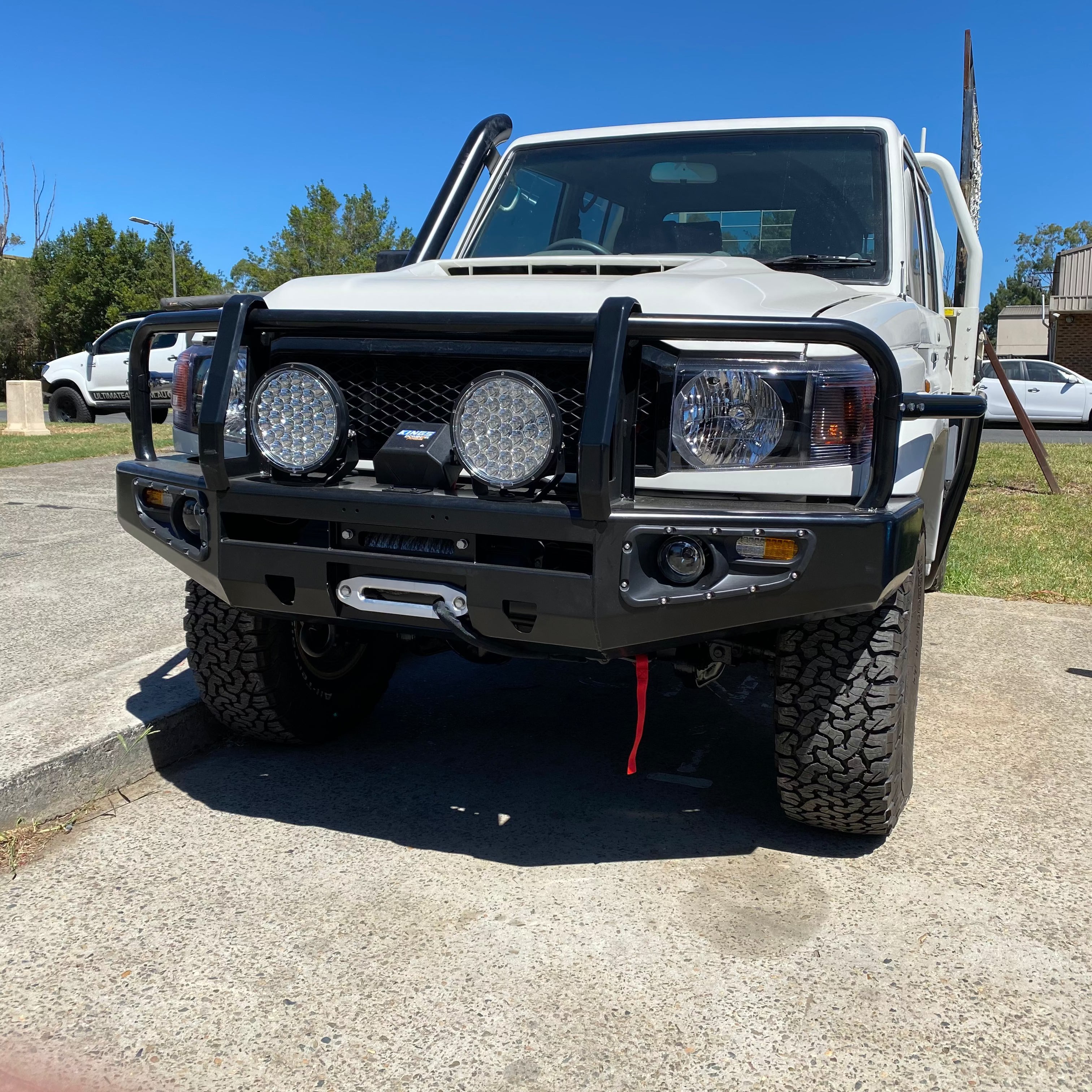 A frontal view of a white off-road vehicle, likely a Toyota Land Cruiser 70 Series, fitted with a heavy-duty black steel bull bar. The bull bar features a winch mounted centrally, two large round LED driving lights, and integrates with the vehicle's fender flares. The vehicle also has a snorkel on the passenger side, large all-terrain tires, and appears to be parked outside on a sunny day.