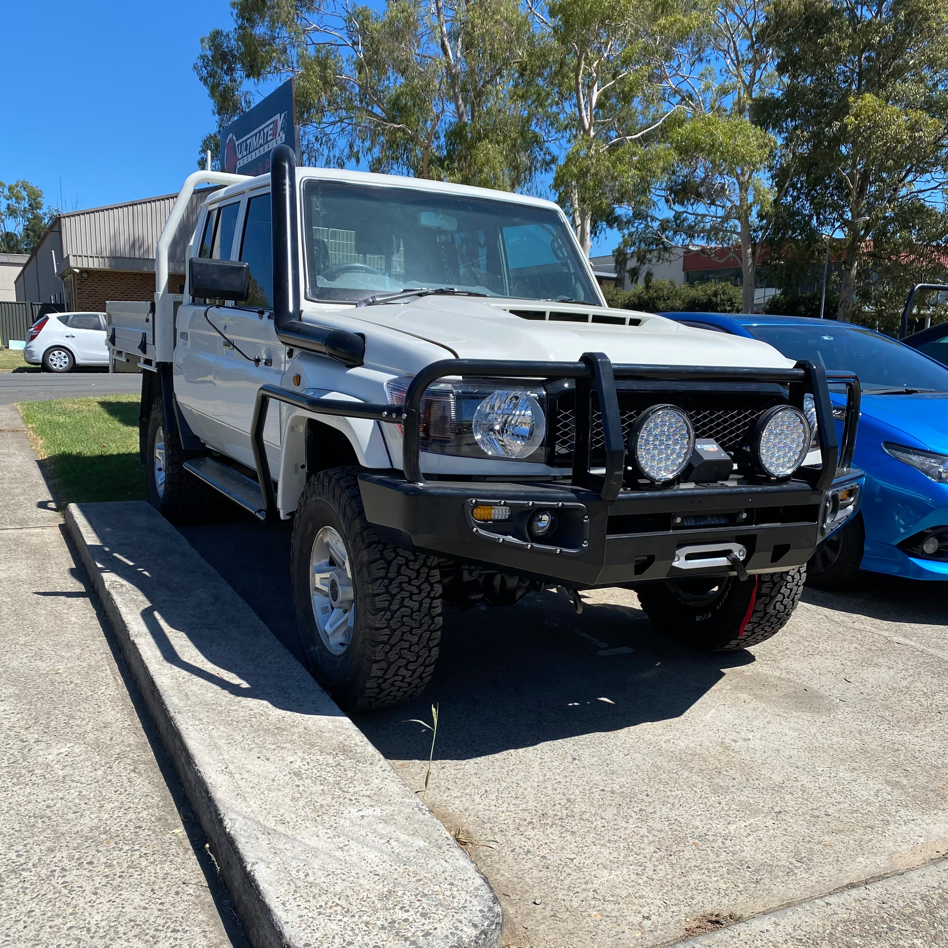 A white Toyota Land Cruiser 79 Series dual cab ute is parked outdoors on a paved lot on a sunny day. The 4x4 is heavily modified with a black steel bull bar, two large round LED spotlights, and a winch visible in the bumper. A black snorkel runs up the driver's side. It has a steel tray back, side steps, and aggressive all-terrain tires on silver alloy wheels. A blue sports car is partially visible on the right, and trees and a building sign are in the background.