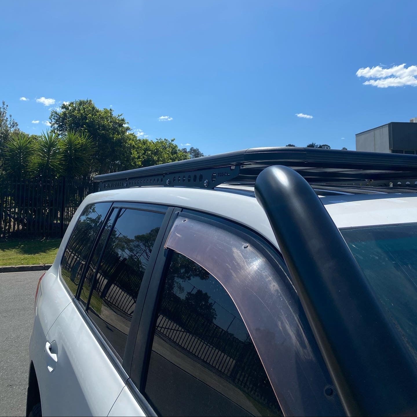 A side-angle photo of a white SUV with several off-road modifications.
Key features visible include a large, flat, black roof rack mounted over the entire roof, and a prominent black snorkel running up the A-pillar on the driver's side. The car also has tinted windows and what appears to be weather shields (rain guards) over the side windows. The vehicle is parked outdoors on a sunny day with trees and a fence in the background.