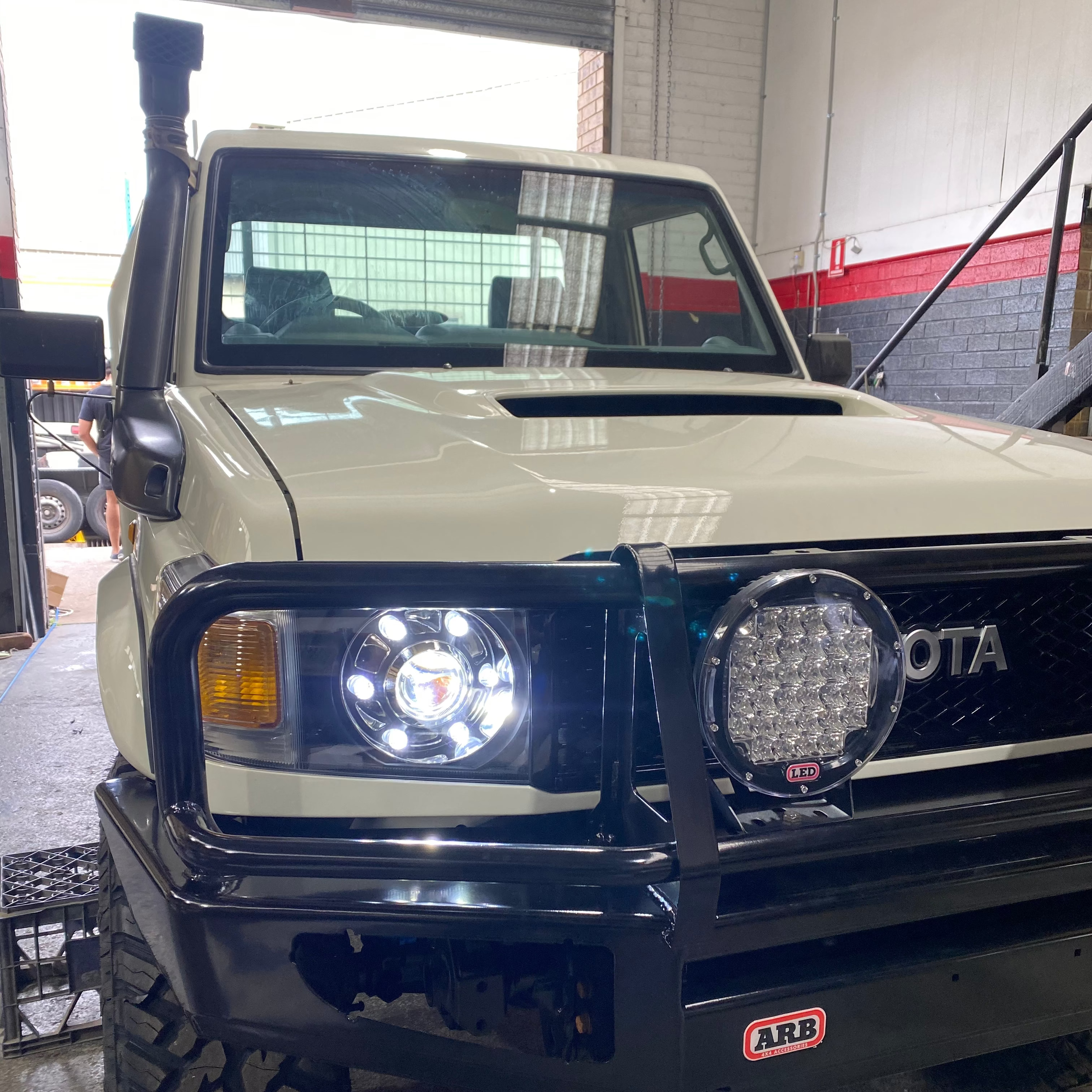 A medium shot of the front end of a white Toyota Land Cruiser 70 Series (or similar 4x4) modified for off-roading, parked inside a garage or workshop.
The vehicle is equipped with a black steel bull bar/bumper that has an ARB logo sticker.
Mounted on the bull bar is a large, round LED driving light with multiple visible LEDs.
The original headlight has been upgraded to a bright, round LED projection headlight featuring a distinct 'halo' or 'angel eye' ring.
An air intake snorkel is visible on the left.