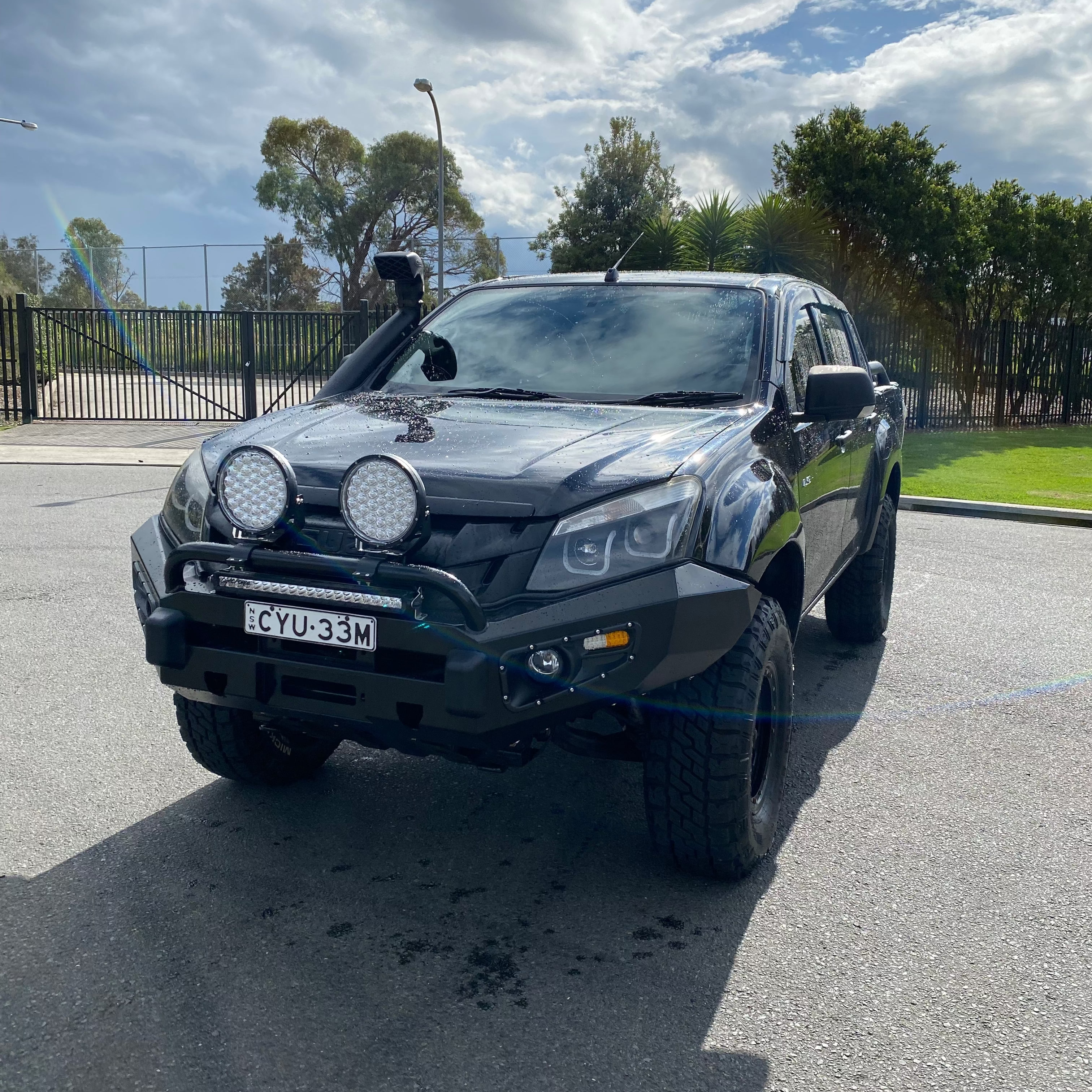 A black, lifted Isuzu D-Max pickup truck, modified for off-roading, is parked outdoors on a paved surface. It is equipped with a black heavy-duty steel bull bar, two large round LED driving lights, a rectangular LED light bar, a snorkel on the driver's side, and large all-terrain tires. A license plate reading "CYU-33M" is mounted on the bumper. In the background, there is a chain-link fence and some foliage.
