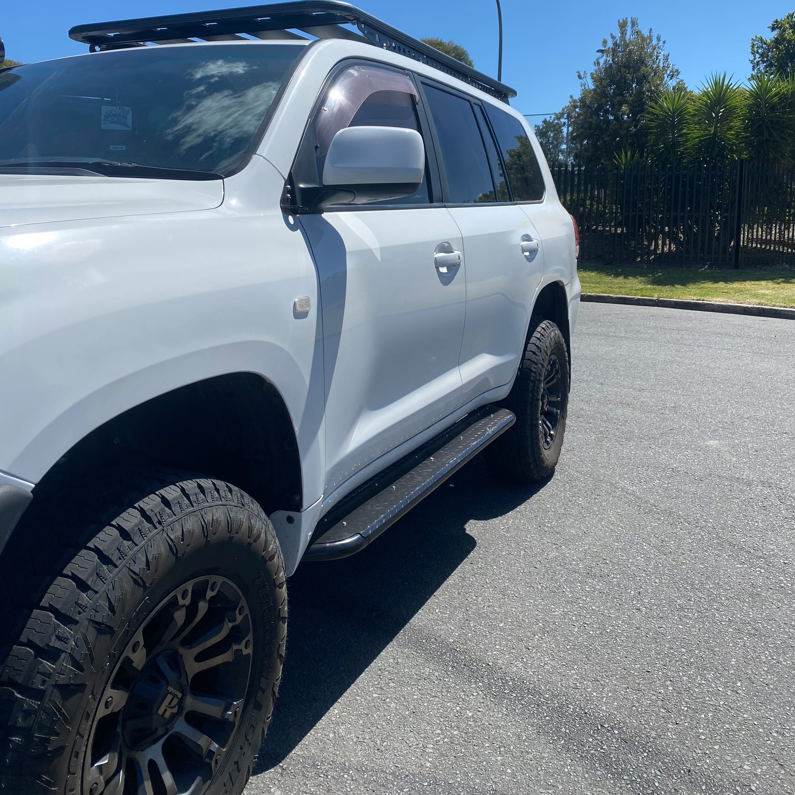 A close-up side view of a white SUV (likely a Toyota Land Cruiser 200 Series) parked on asphalt under bright sunlight. The image highlights the vehicle's modifications, including large, aggressive off-road tires mounted on black aftermarket wheels, a black rock slider/side step, and a platform roof rack visible on the top. The background features a curb, some green foliage, and a fence. The photo captures the rugged build and off-road readiness of the vehicle.