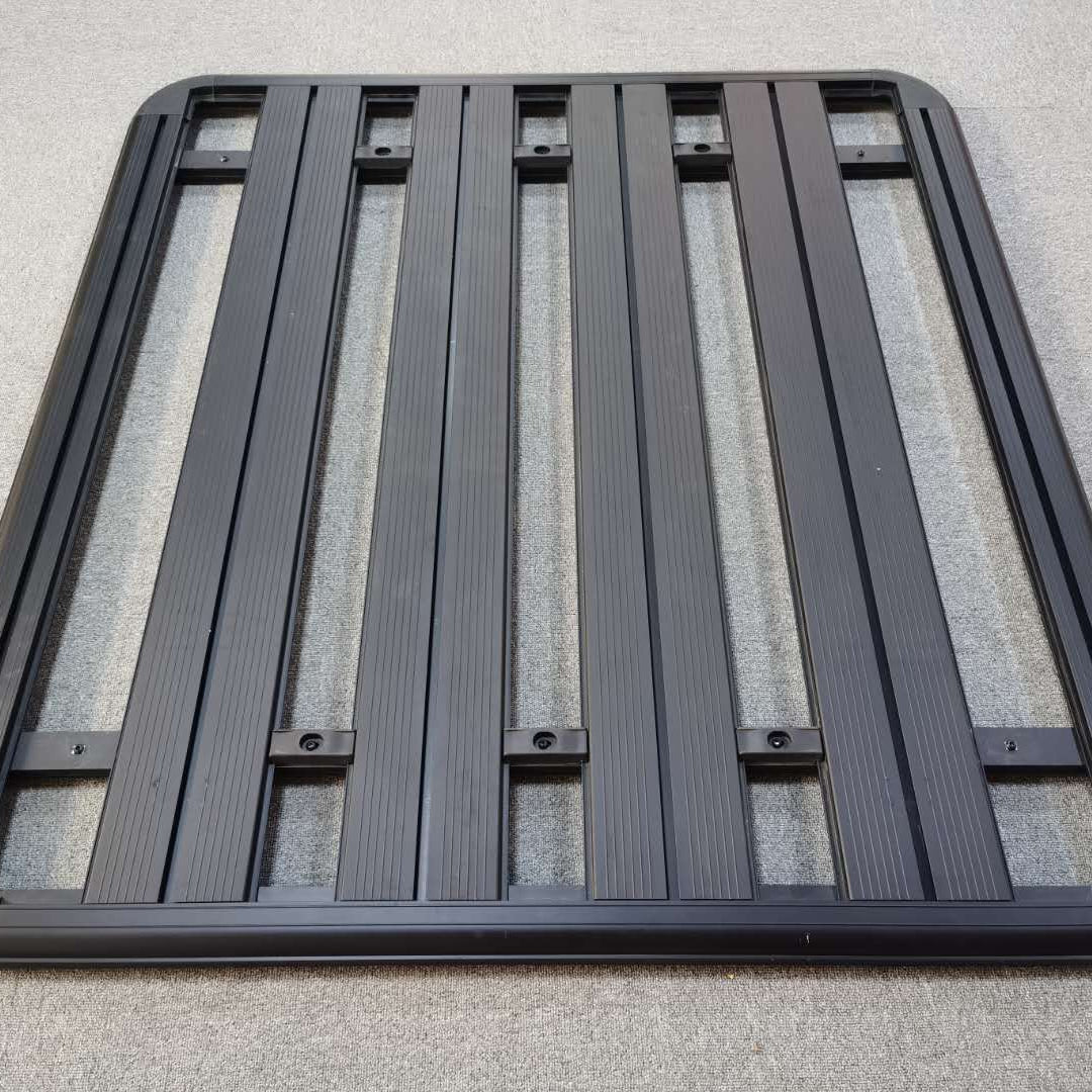 A close-up, overhead view of a new, black, rectangular flat roof rack platform.
The rack is constructed of aluminum and features multiple parallel slats or planks running lengthwise, secured within a heavy-duty frame. The slats have grooves for mounting accessories. The rack is designed for an SUV or truck and is resting on a gray, carpeted surface.
