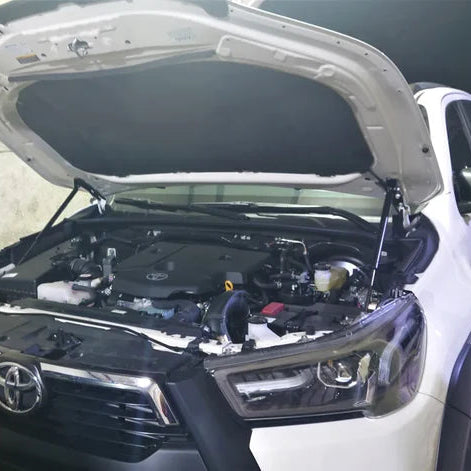 
A white pickup truck, which appears to be a Toyota Hilux, is shown with its hood open. The engine bay is visible, and the hood is held up by a pair of aftermarket or custom-installed hydraulic struts (gas springs) instead of a traditional prop rod. The truck is parked indoors or in a garage.