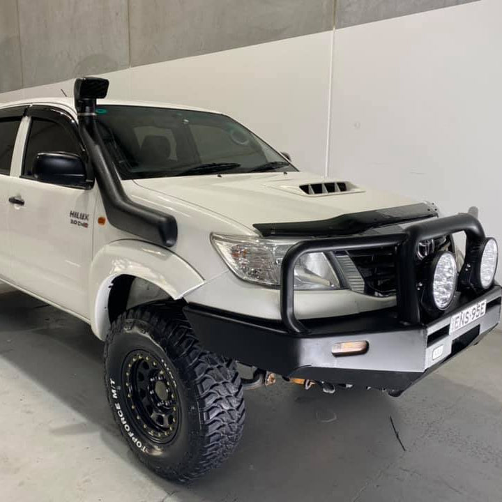 A front three-quarter view of a heavily modified white Toyota Hilux pickup truck, parked indoors in what looks like a garage or showroom. The vehicle features a large black snorkel mounted on the driver's side, a heavy-duty black steel bull bar with two large auxiliary spotlights, black off-road wheels with large knobby tires, and a dark plastic hood protector/deflector.