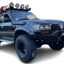 Close-up of the front right quarter of a dark green or black Toyota Land Cruiser (likely 80-Series) equipped for off-roading, featuring a snorkel, heavy-duty aftermarket bumper, large off-road tires on black wheels, fender flares with exposed rivets, auxiliary lights on the roof rack, and a round auxiliary light mounted on the bumper. The background is a checkered transparency pattern.