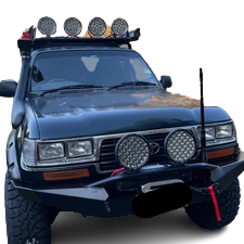 Front view of a dark green, heavily modified off-road vehicle, likely a Toyota Land Cruiser 80-Series. The vehicle features a large snorkel on the driver's side, a heavy-duty steel front bumper, an antenna mounted on the passenger side, two large round LED auxiliary lights mounted on the bumper, and four smaller round LED lights mounted on a roof rack. The license plate area on the bumper is redacted. The background is a checkered transparency pattern.