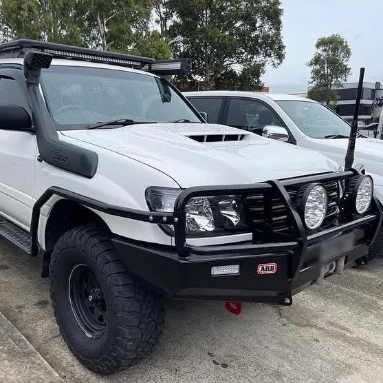 
A white, off-road modified SUV (likely a Toyota Land Cruiser) is parked outside. It features several aftermarket accessories, including a large, black snorkel on the driver's side, an aggressive black steel bull bar with an integrated winch mount and two large driving lights, a small LED light bar mounted on the bull bar, and a hood scoop. The vehicle has large, knobby off-road tires on black wheels and a roof rack with an overhead LED light bar. Another white vehicle is partially visible behind it.
