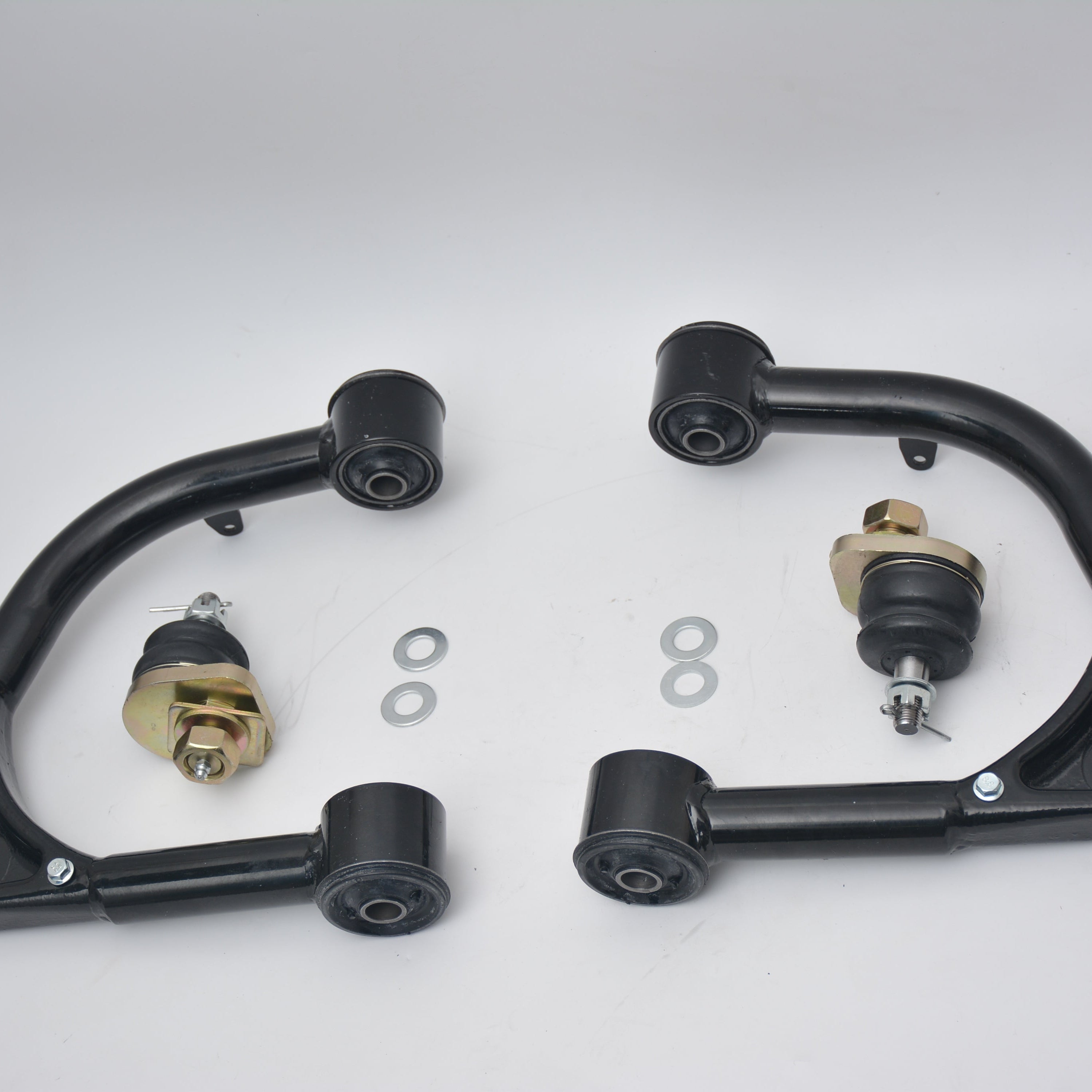 Two black fabricated upper control arms featuring pre-installed bushings, paired with two uninstalled gold-colored ball joints and four metal washers on a white background.