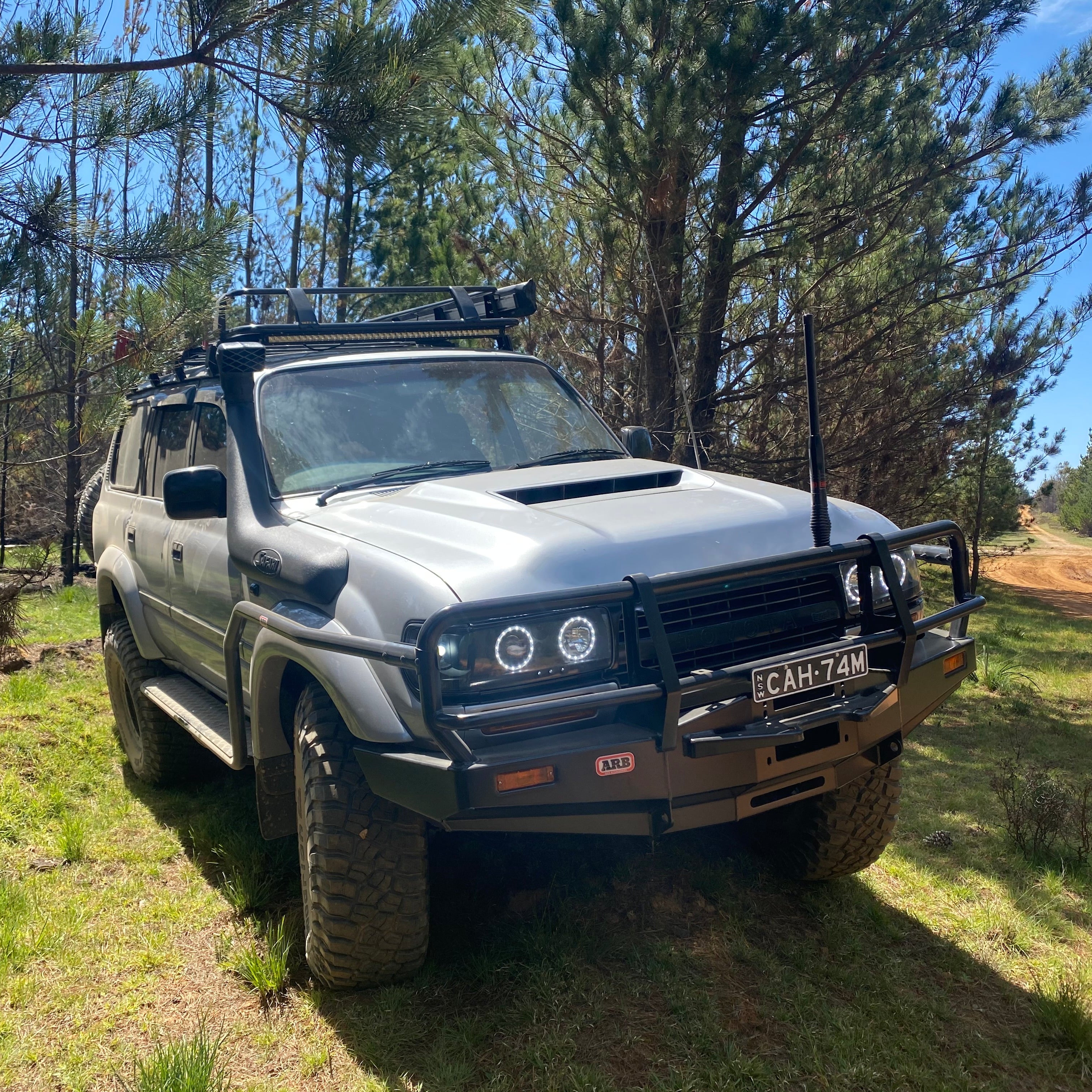 A rugged off-road vehicle (Toyota Land Cruiser 80 Series) with numerous modifications sits ready for adventure on a grassy trail surrounded by pine trees, with bright sunlight highlighting the front of the truck.