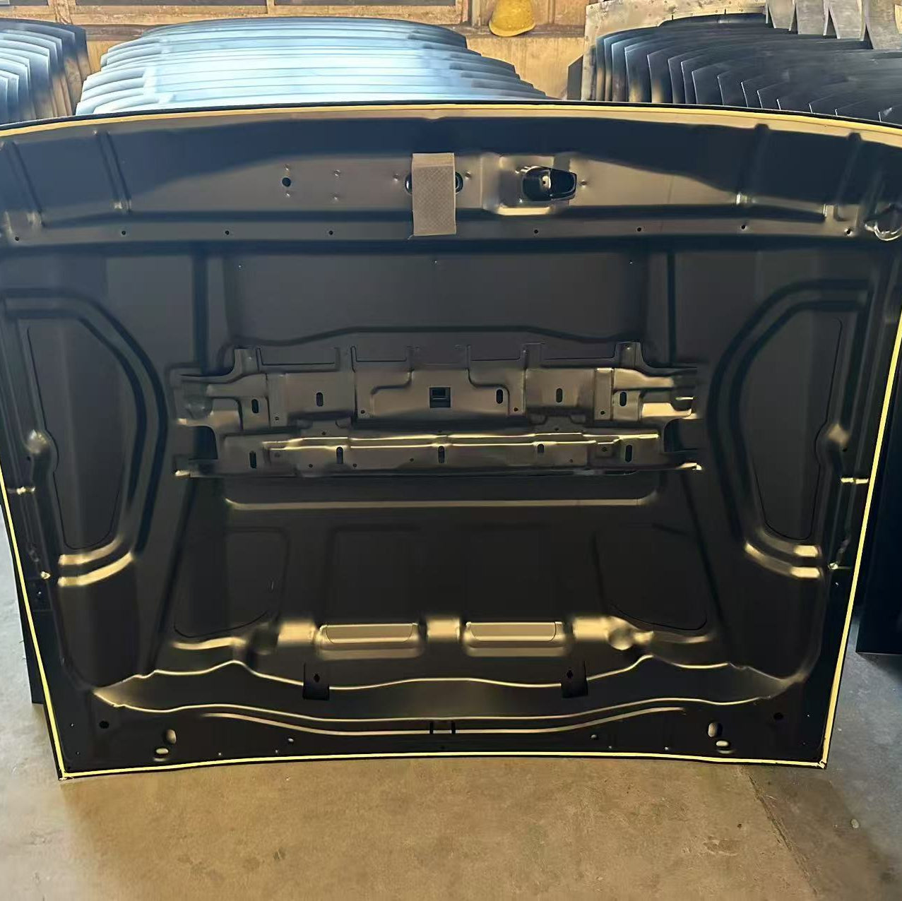 
A photograph showing the underside of a black car hood with a reinforced metal frame structure. The underside is painted black, and the edges show a white or light trim, likely the unpainted substrate. It is being stored in a warehouse or facility, and a stack of similar car hoods is visible in the background. The hood features mounting points and a central support bracing.