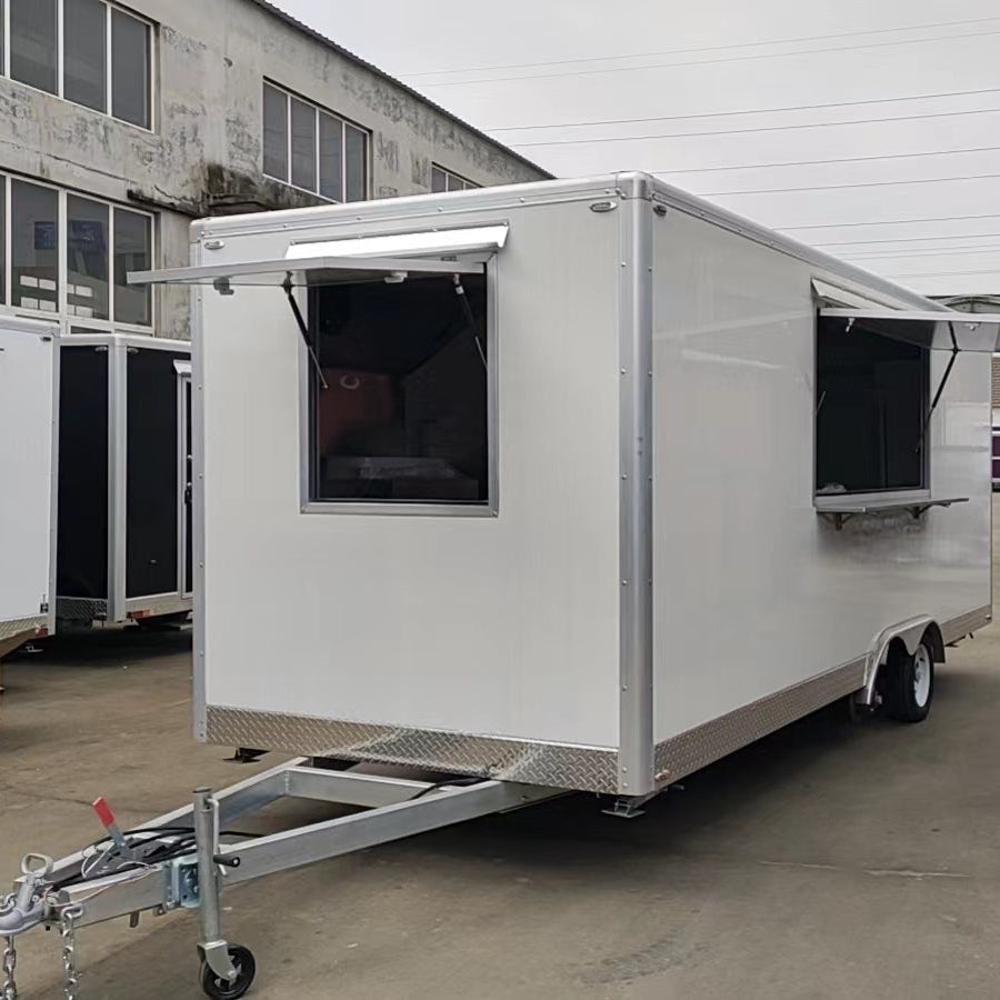 A photograph of a white, rectangular mobile food concession trailer parked outdoors, likely in a lot or near a warehouse. The trailer has a silver-colored metal trim and two large service windows on the side facing the viewer. Both windows have awning-style openings that are propped open, and the right-hand window features an exterior serving ledge. The trailer is hitched to a silver-colored A-frame tongue with a single wheel set (axle not fully visible). In the background, there is a large, aging building