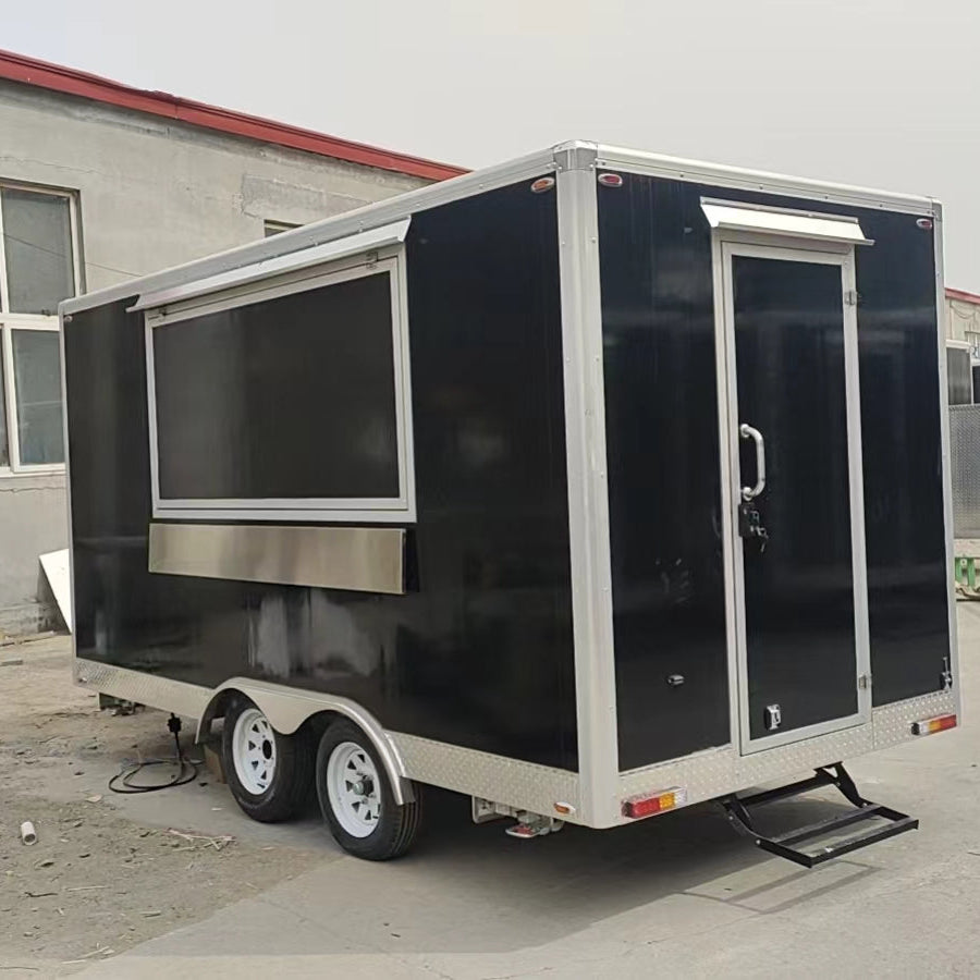 A three-quarter view photo of a black enclosed trailer, likely a mobile food or concession unit, parked outdoors on a paved surface. The trailer has a large service window on the left side, which is currently closed and covered by a white awning or hatch at the top. Below the window, there is a horizontal silver metallic counter space. The back right of the trailer features a single white door with a silver handle, and a retractable metal step is extended below the door. The bottom perimeter of the trailer